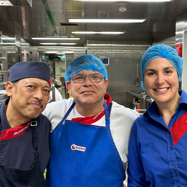 Três profissionais da Sodexo trabalhando na área de Offshore, usando uniformes e toucas de proteção, sorrindo e mostrando espírito de equipe na cozinha industrial.