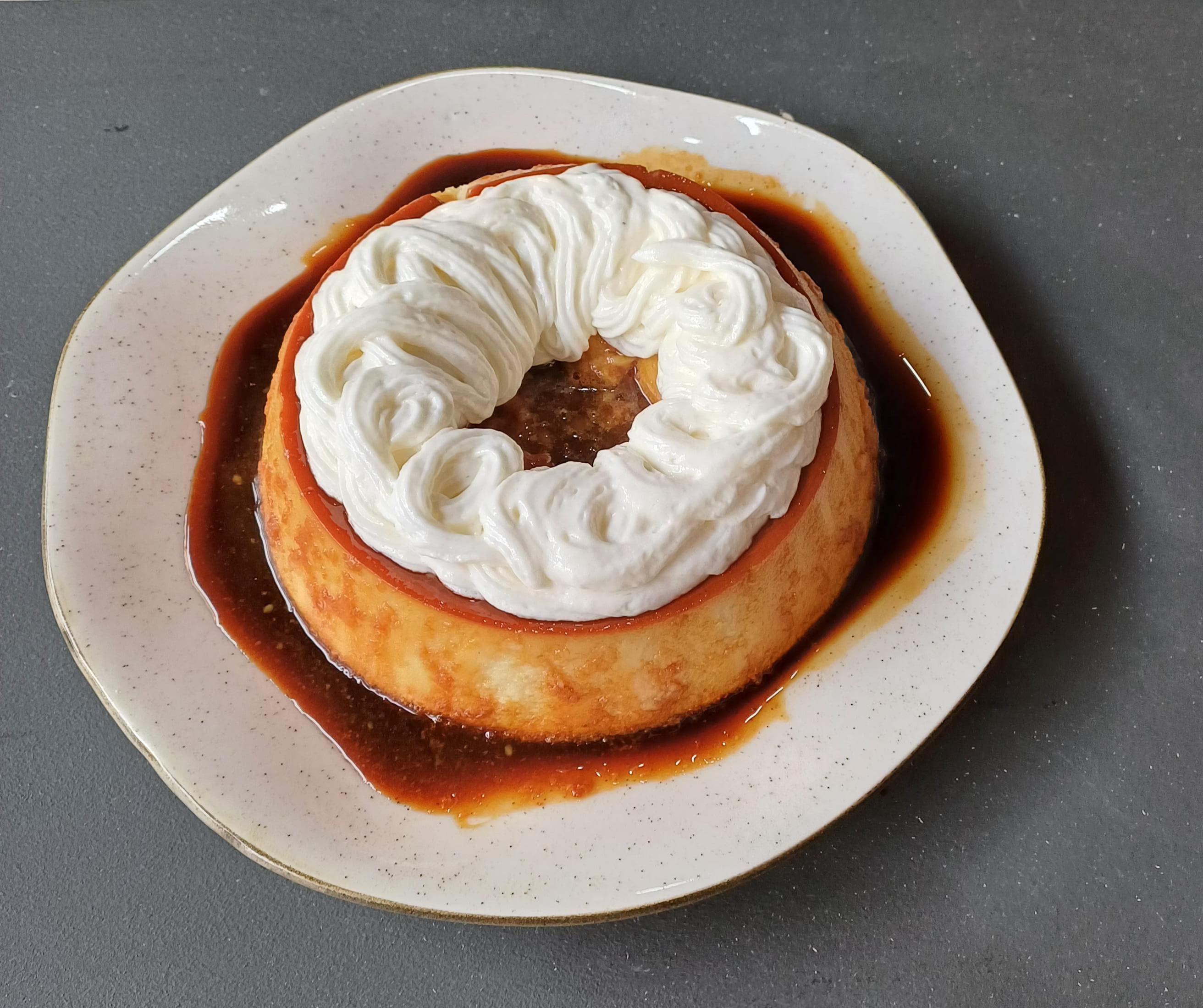 Pudim de leite com calda de café servido em prato claro, decorado no topo com chantilly em formato espiral, com a calda escura escorrendo ao redor da sobremesa. 