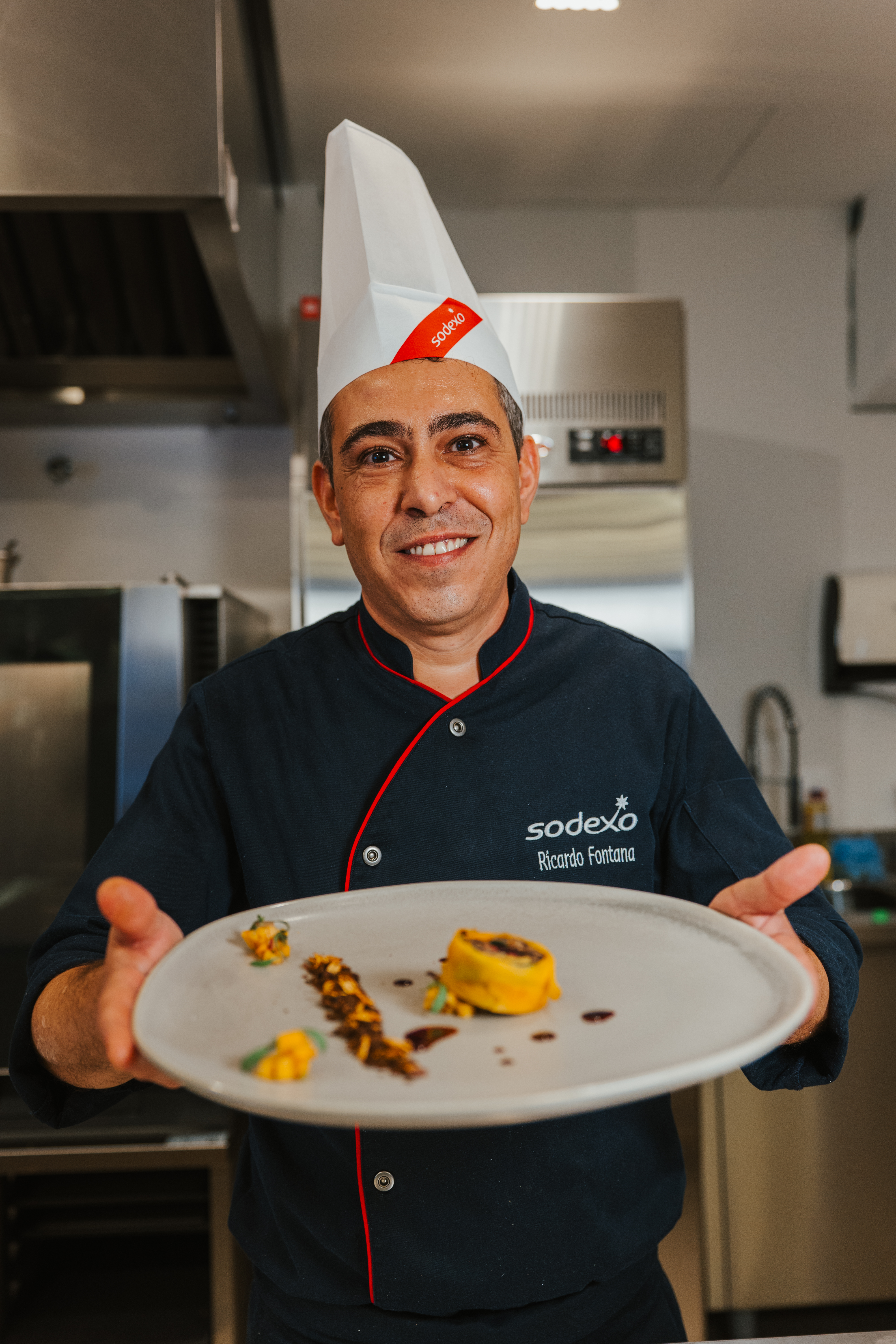 Chef da Sodexo, Ricardo Fontana, sorrindo enquanto segura um prato gourmet montado, usando uniforme azul marinho e touca branca, em uma cozinha profissional.