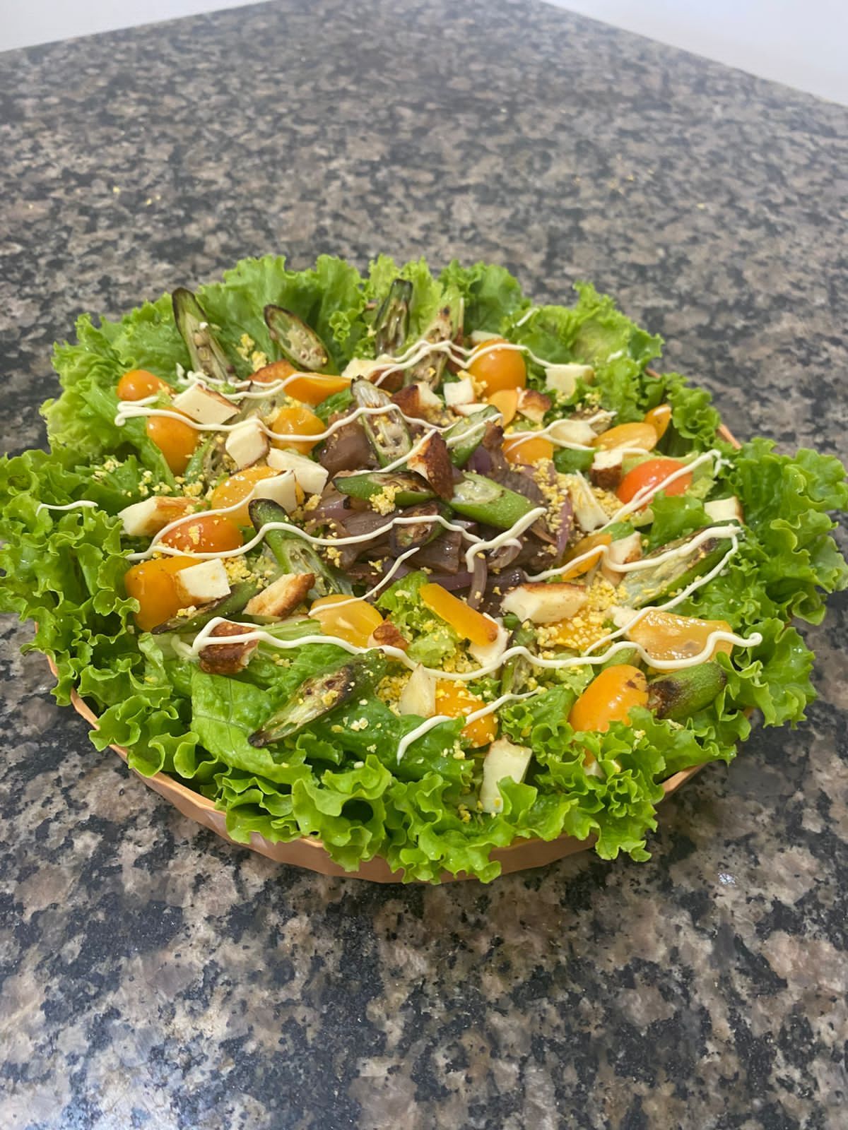  Salada montada em travessa redonda com folhas de alface crespa, carne de sol desfiada, quiabo tostado, cubos de queijo coalho, tomates amarelos, croutons de cuscuz e fio de molho por cima.