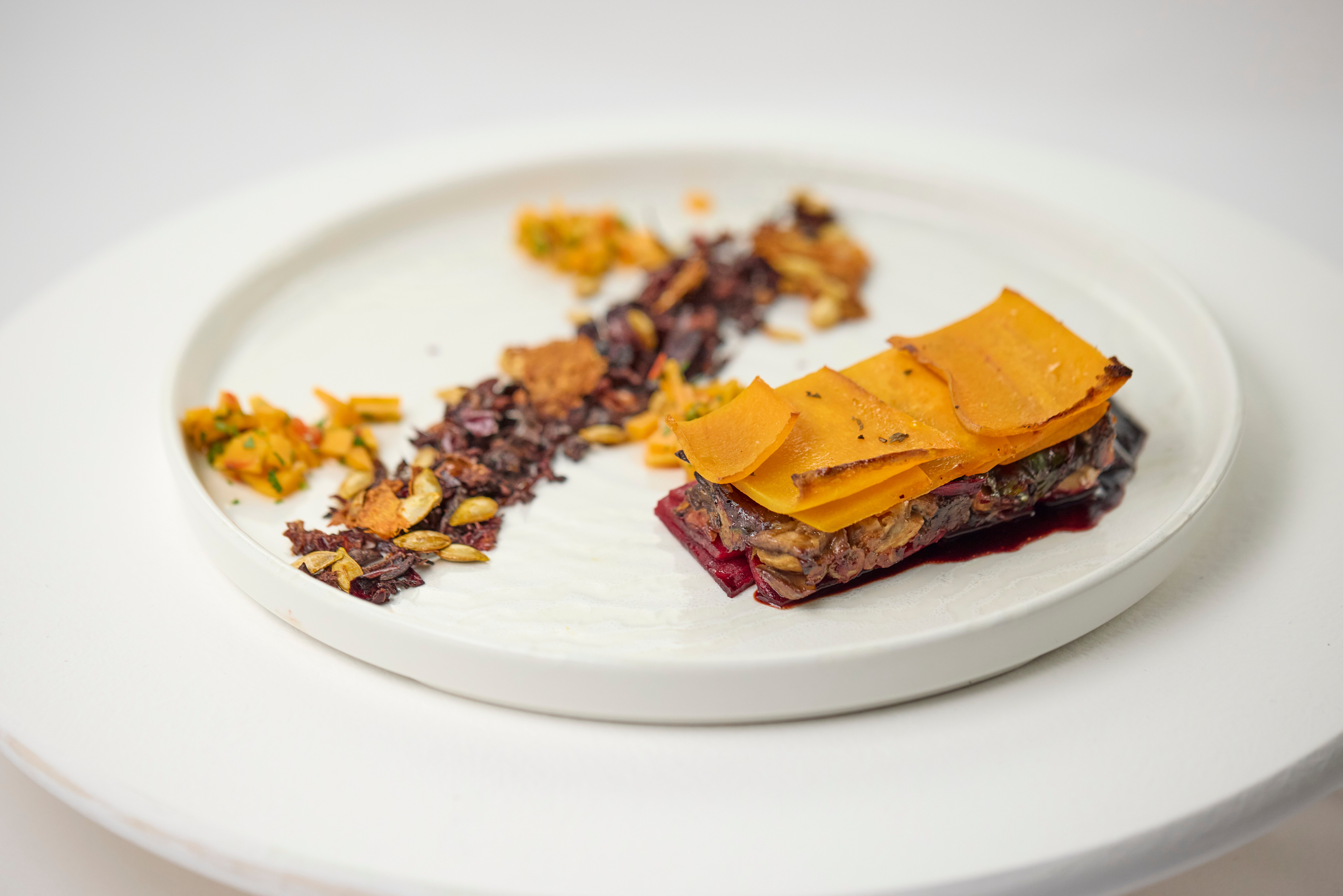  Prato sofisticado com montagem contemporânea: camadas de legumes e grãos moldados em formato retangular, cobertas por lâminas finas de abóbora. Ao lado, elementos crocantes, sementes e pequenos acompanhamentos coloridos dispostos artisticamente em prato branco. 