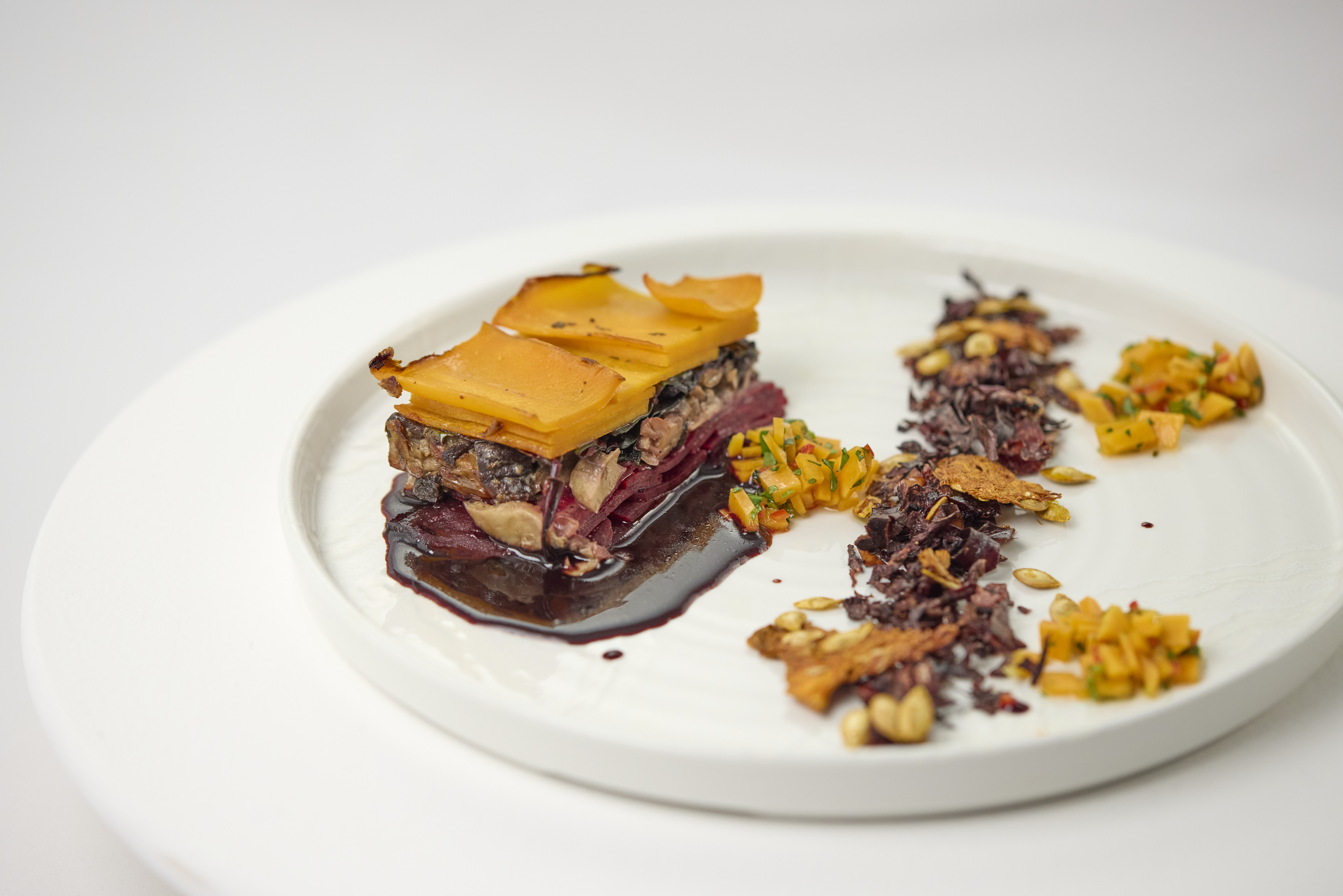 Prato gastronômico com apresentação refinada: terrine de legumes e grãos em camadas, coberta por lâminas finas douradas, servida sobre molho escuro. Ao redor, acompanhamentos dispostos de forma artística, com sementes, farofa crocante e pequenos cubos de legumes coloridos em prato branco.