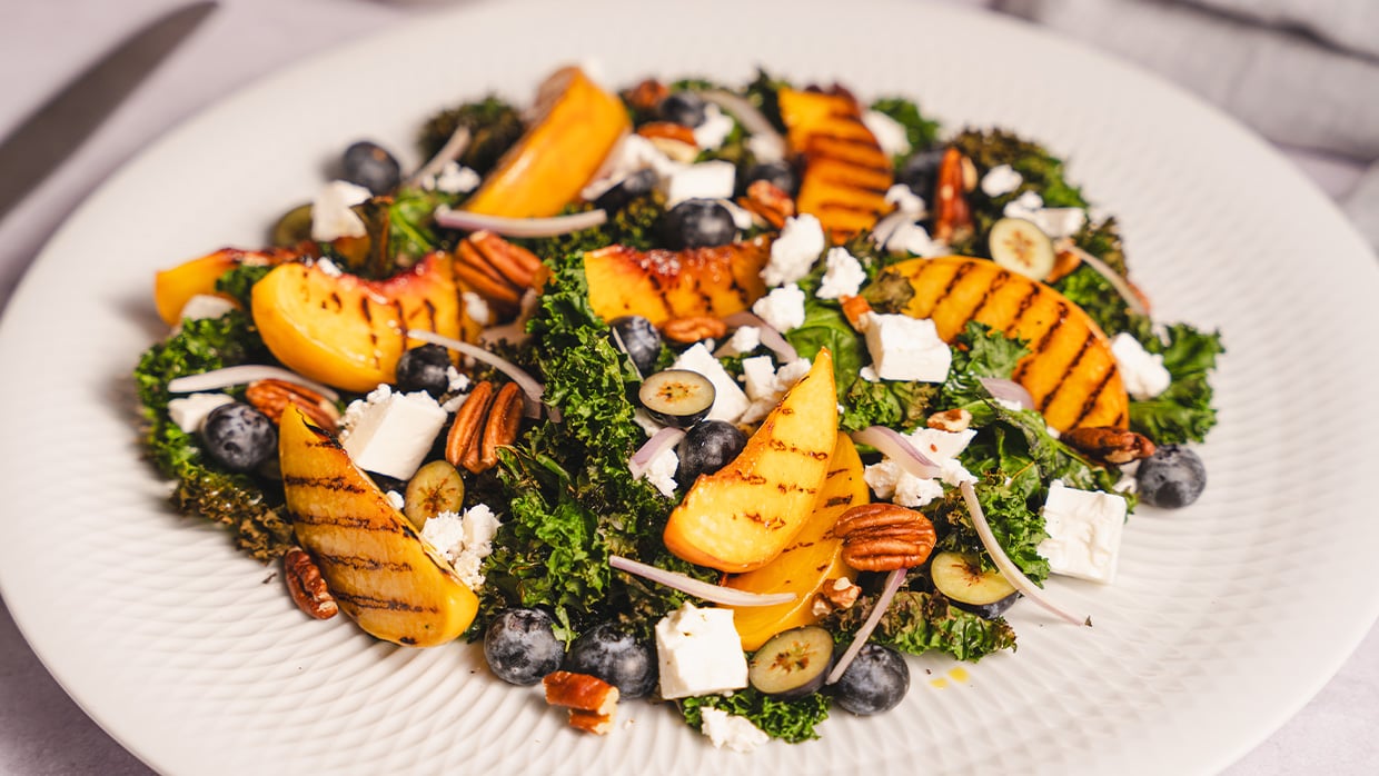 Prato de salada com folhas verdes (couve), fatias de pêssego grelhado, mirtilos, nozes-pecã, cubos de queijo branco e cebola roxa, finalizada com molho tipo vinagrete, servida em prato branco.