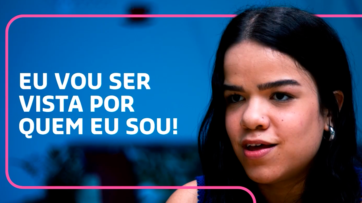 Mulher de cabelos longos e pretos, com uma expressão determinada, aparece ao lado de uma frase motivacional: "Eu vou ser vista por quem eu sou!". O fundo é azul e a imagem está enquadrada com uma linha rosa, destacando o texto e a mensagem.