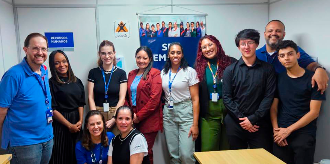 Equipe da Sodexo reunida em ambiente corporativo, posando para foto em uma sala com identificação de Recursos Humanos ao fundo. O grupo é composto por profissionais diversos, usando crachás, com postura sorridente e colaborativa, representando o time responsável pelas operações e gestão de pessoas.
