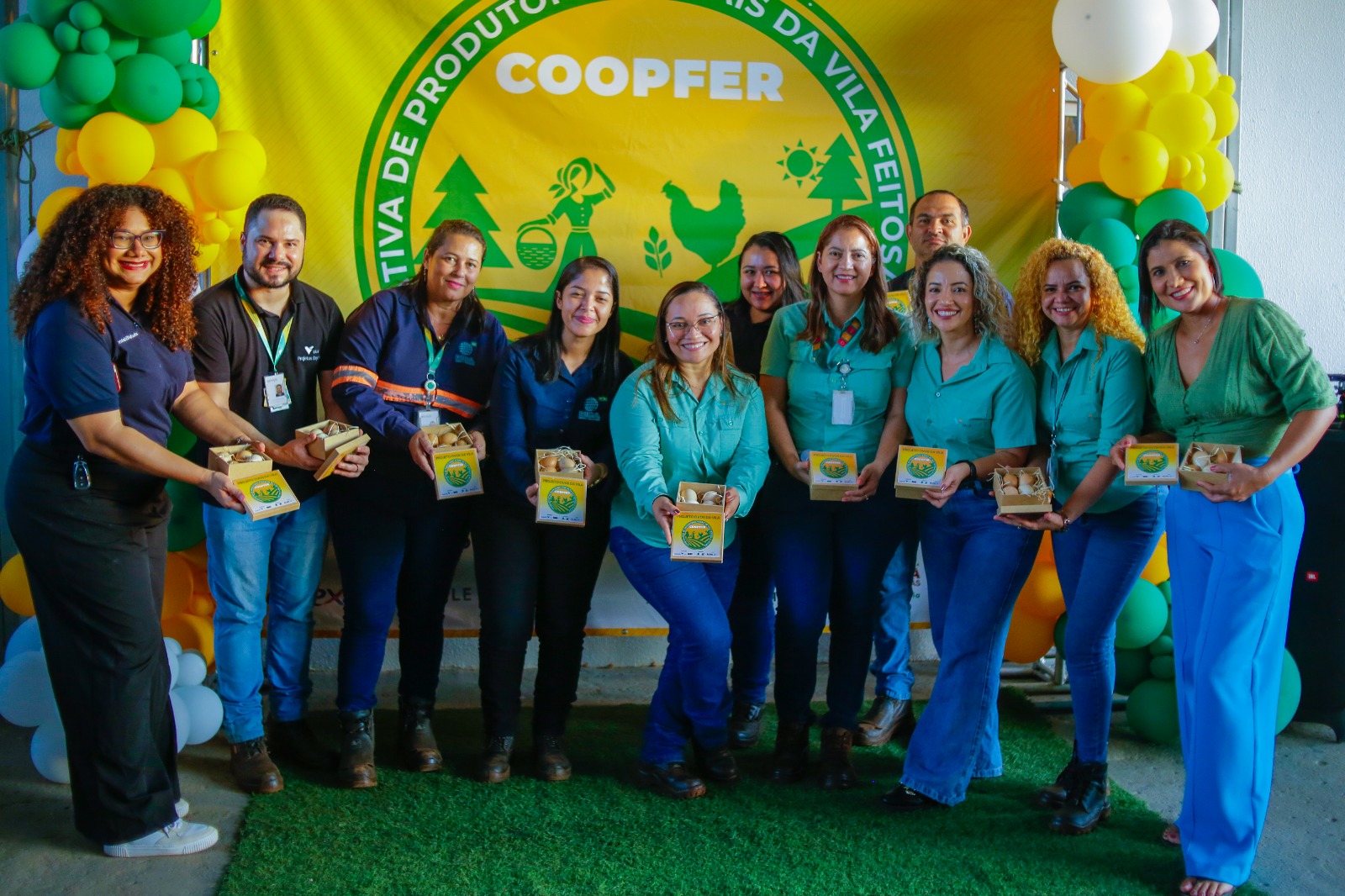 Grupo de mulheres e homens posa sorrindo segurando caixas de ovos caipiras em frente ao banner da cooperativa COOPFER, decorado com balões verdes e amarelos.