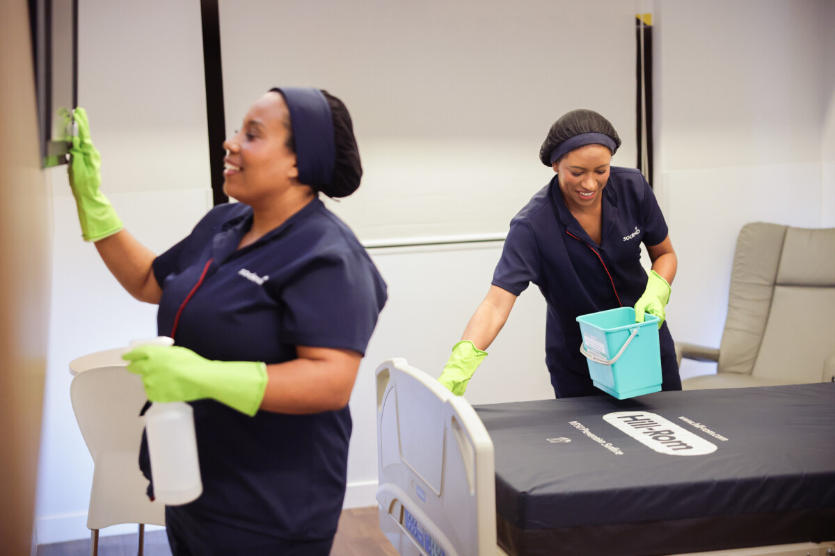 Colaboradoras da Sodexo realizam limpeza hospitalar, utilizando luvas e produtos de higienização, em um ambiente interno com leito e poltrona. 