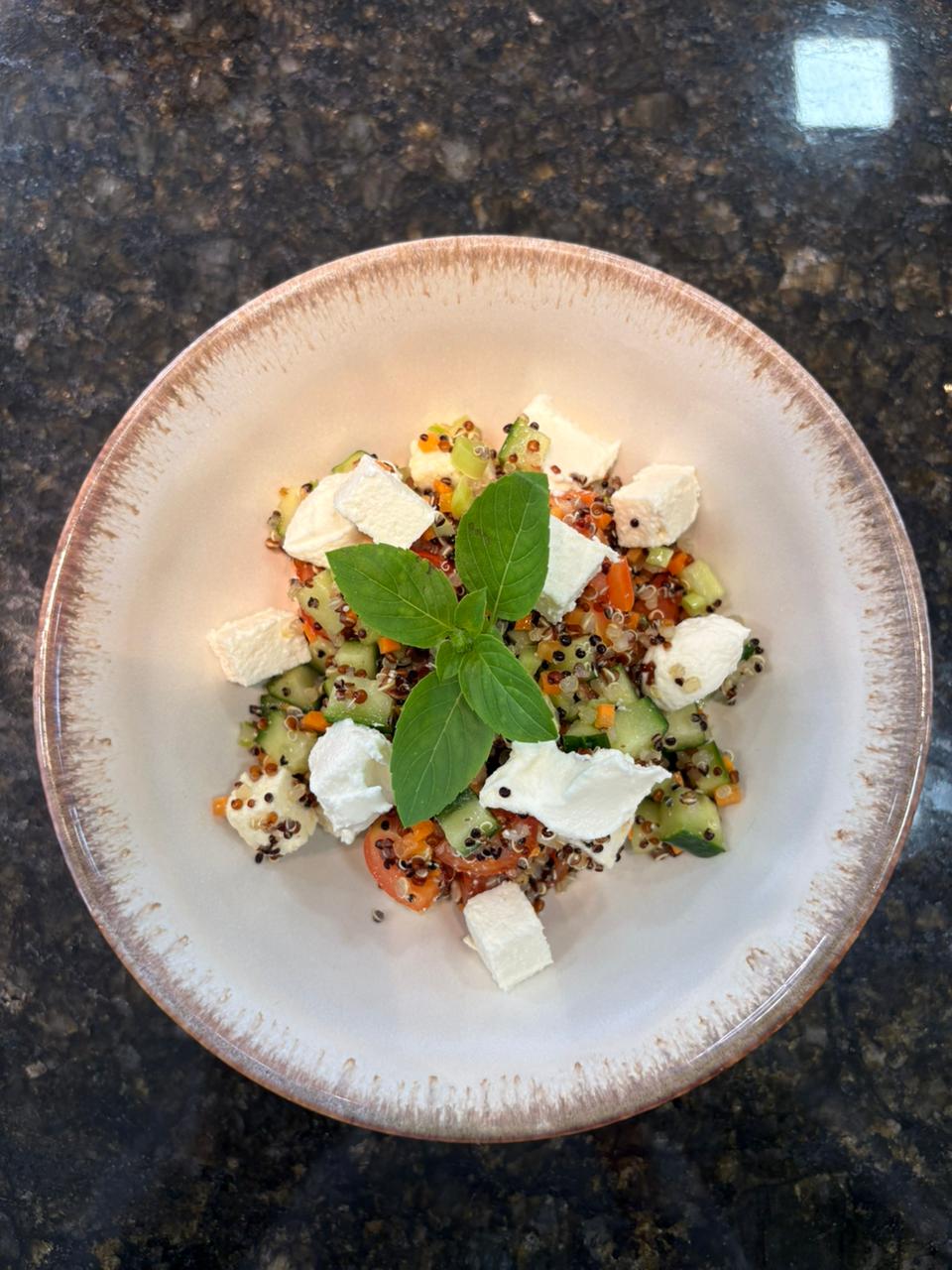  Bowl visto de cima com salada de quinoa, cubos de pepino e tomate, finalizada com pedaços de ricota branca e folhas frescas no centro, sobre bancada escura.