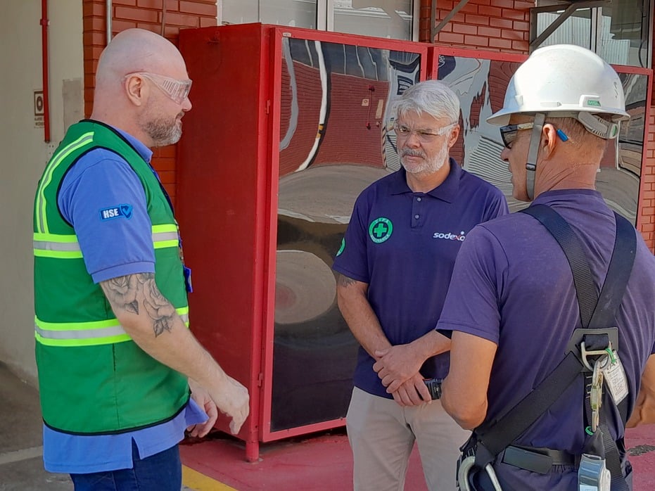  Três profissionais em área industrial conversam em frente a um equipamento de incêndio. Dois usam uniforme da Sodexo, com capacete, óculos de proteção e EPI, enquanto o terceiro veste colete verde de segurança (HSE). 