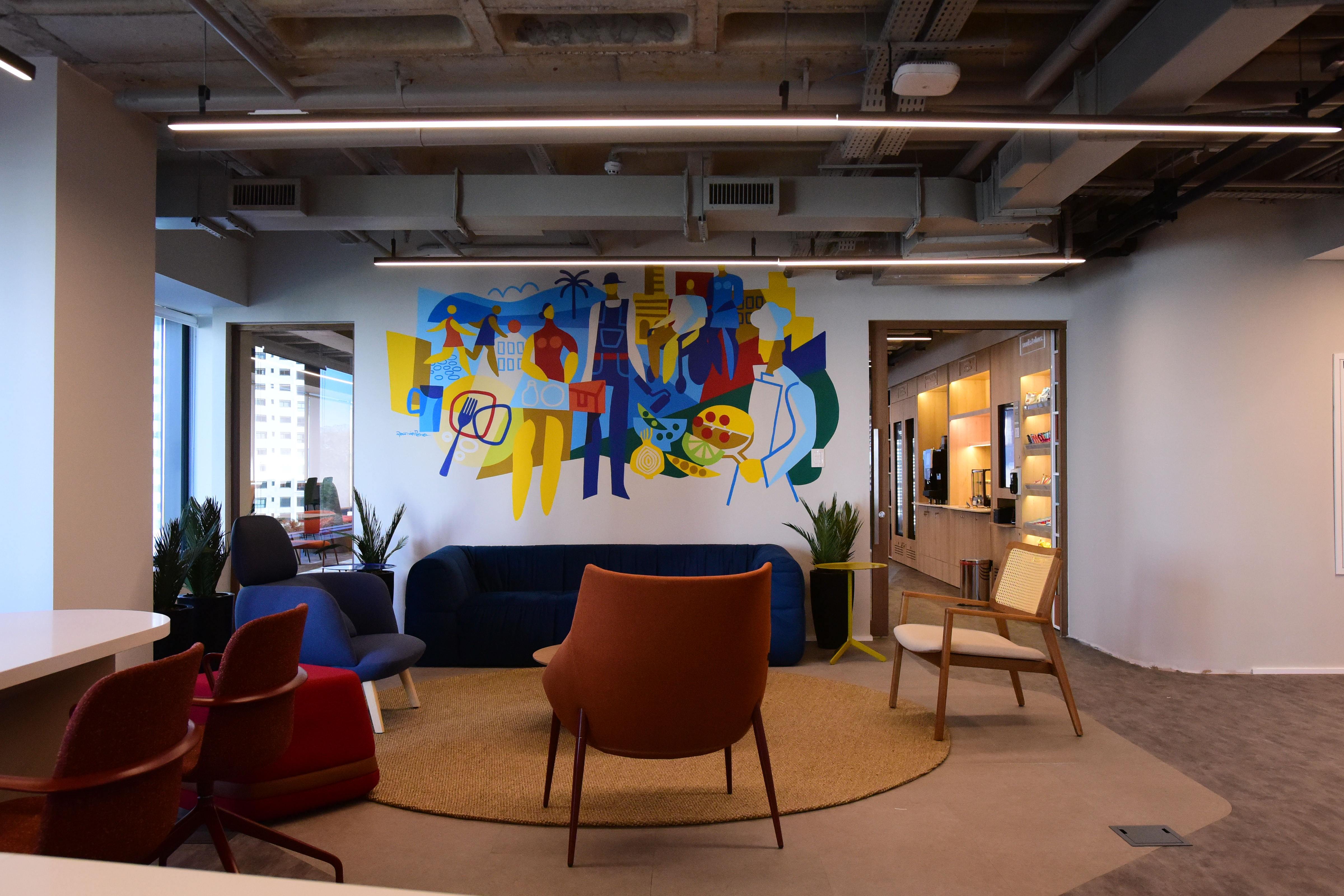 Espaço interno moderno com área de convivência, poltronas e sofás coloridos, plantas e um mural ilustrado com figuras estilizadas e elementos ligados à alimentação e à cultura brasileira na parede ao fundo.