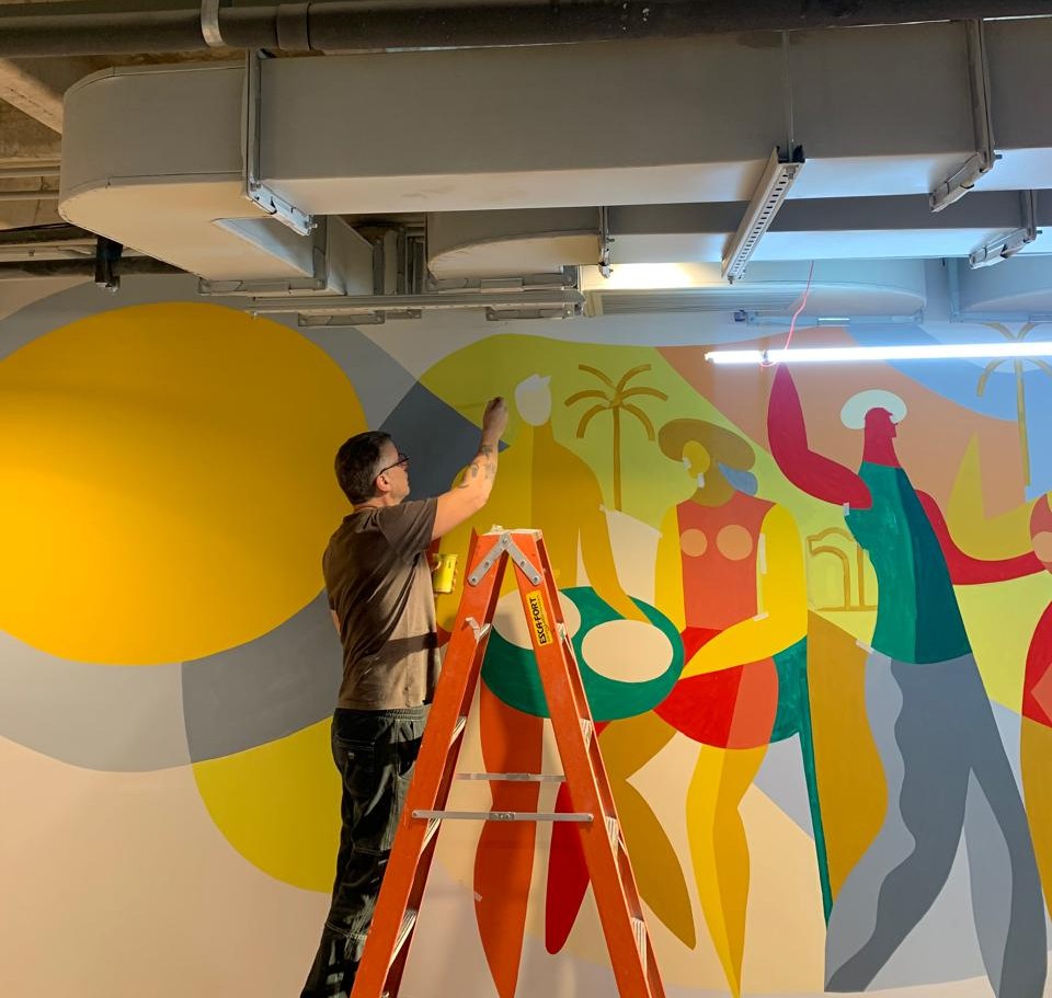  Artista em pé sobre uma escada pintando um mural colorido em parede interna, com figuras estilizadas que representam a cultura brasileira. O ambiente é um espaço corporativo com teto aparente, dutos de ar e luminárias, evidenciando o processo de criação da obra.