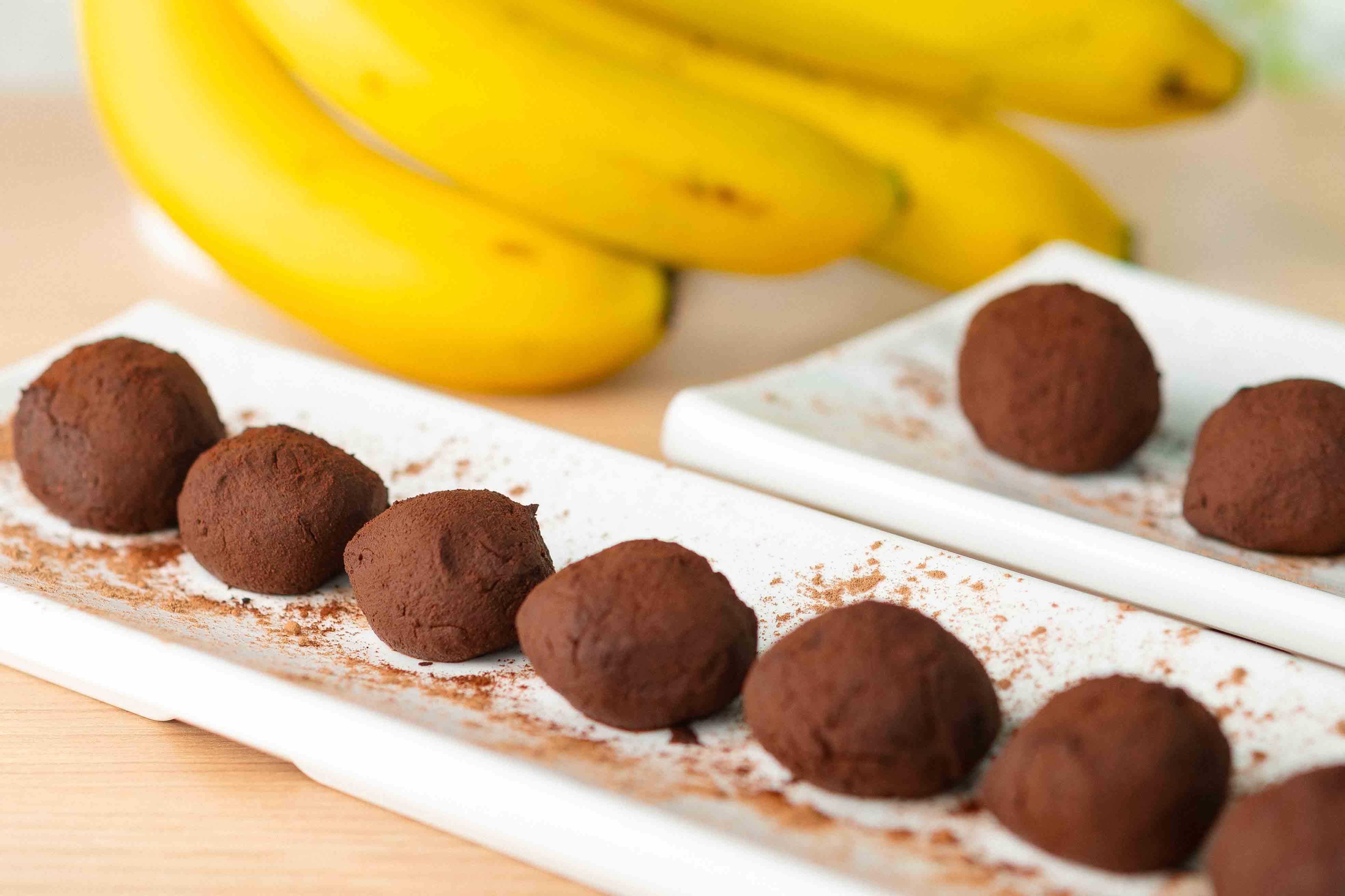 Docinhos de chocolate polvilhados com cacau, dispostos em fileiras sobre travessas brancas, com bananas maduras ao fundo, sobre mesa de madeira clara.