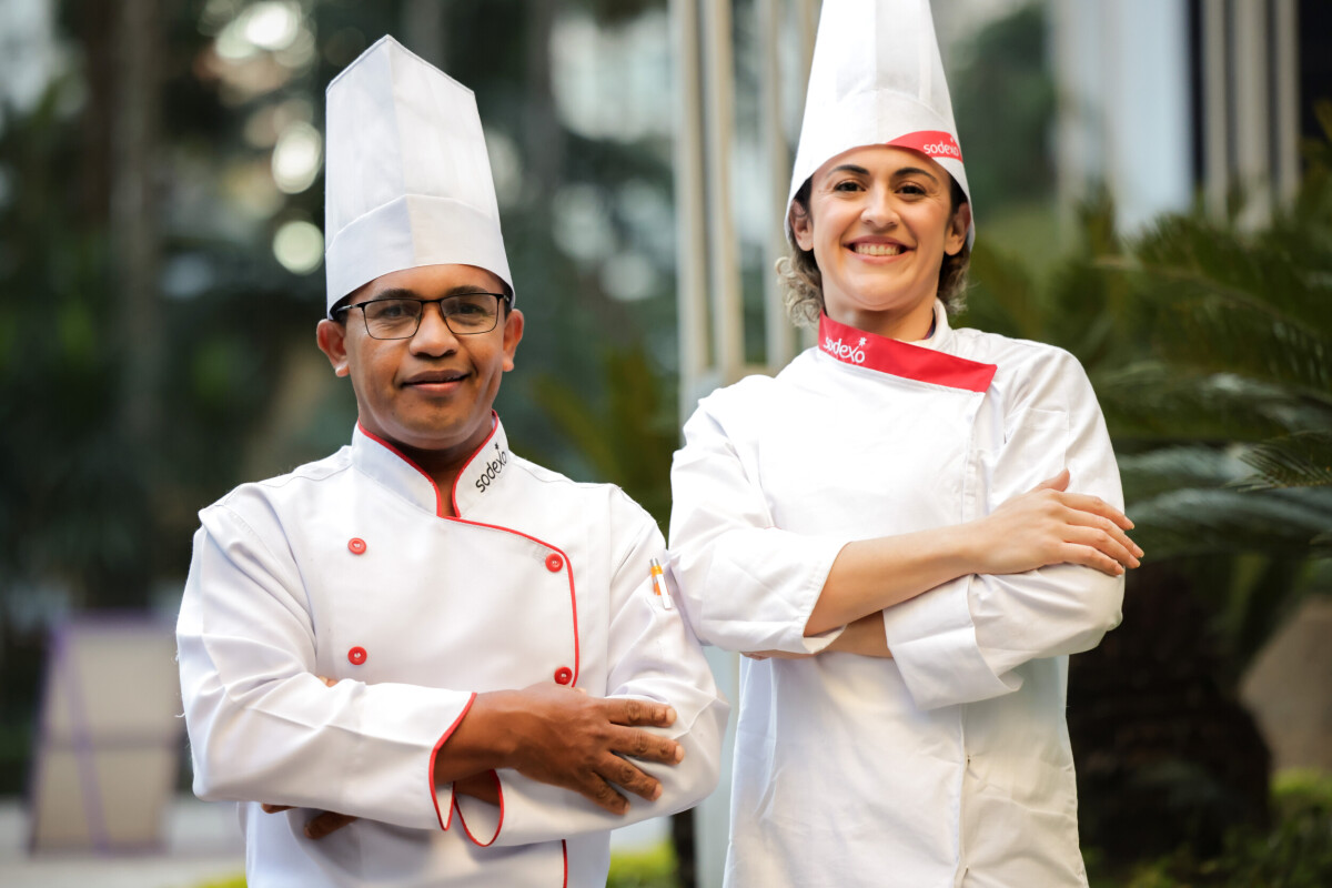 Dois chefs da Sodexo, um homem e uma mulher, vestindo uniformes brancos com detalhes vermelhos e toques da marca, estão lado a lado em ambiente externo, sorrindo para a câmera, com os braços cruzados, transmitindo profissionalismo e orgulho.