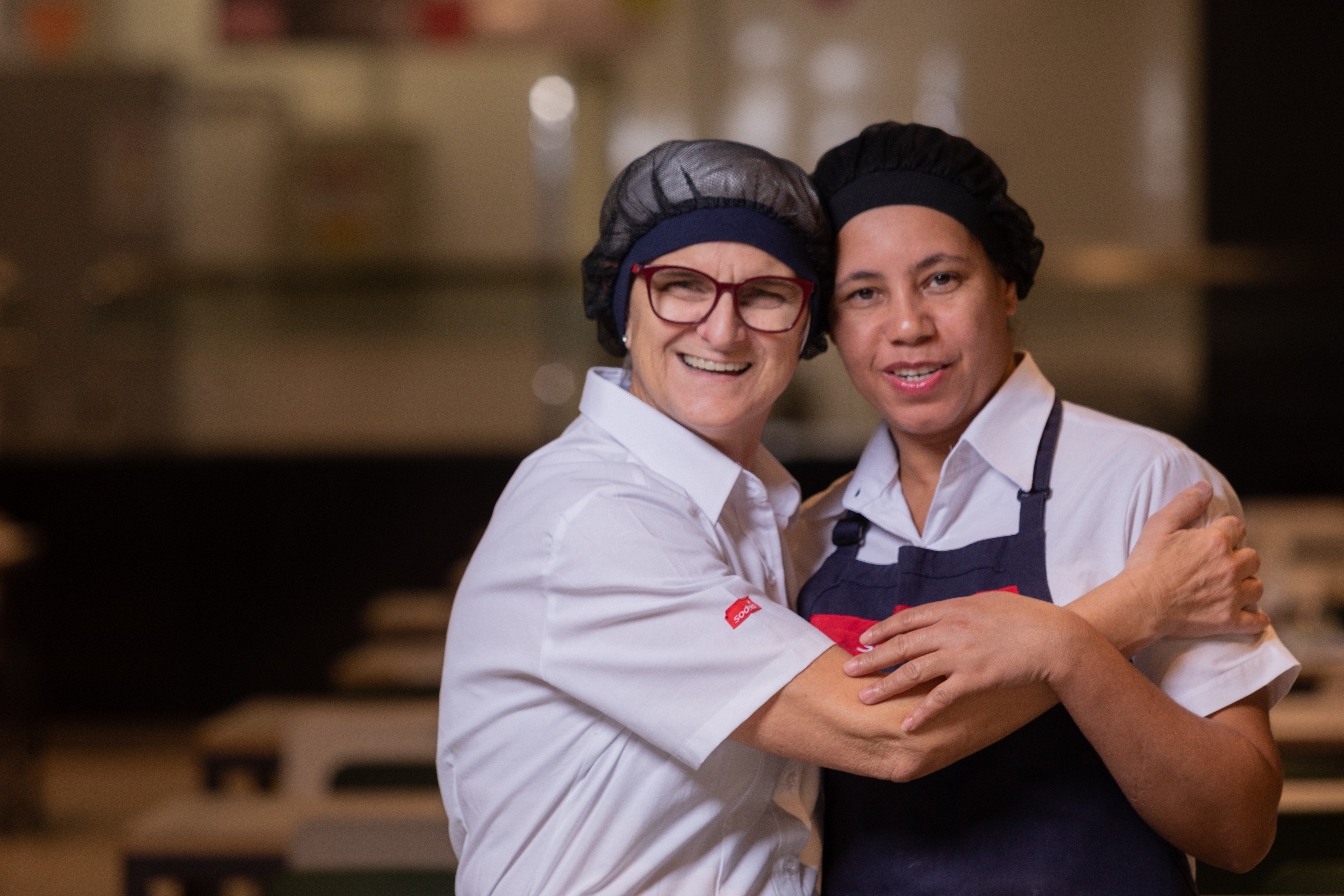 Duas colaboradoras uniformizadas sorriem para a câmera em refeitório
