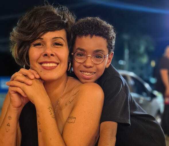  Mulher de cabelo curto sorrindo enquanto abraça um menino de óculos em um momento de carinho e conexão. Os dois estão apoiados sobre uma mesa vermelha em um ambiente externo durante a noite.