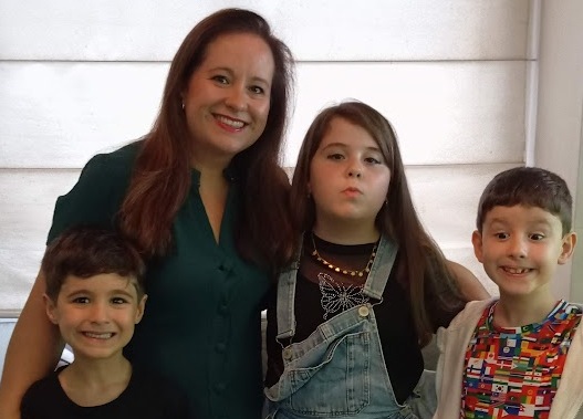  Mulher sorrindo posa ao lado de três crianças em um ambiente interno iluminado. Uma menina e dois meninos estão próximos dela, em um momento de família e carinho. 