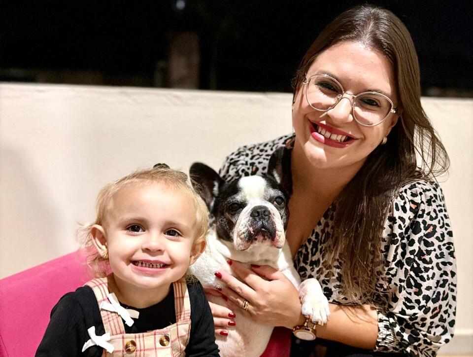  Foto de uma mulher e uma criança sorrindo ao lado de um cachorro da raça bulldog francês. A criança está sentada em uma poltrona rosa, segurando um brinquedo e uma xícara pequena, usando vestido xadrez e blusa preta. 