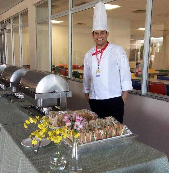 Chef Jairo, uniformizado com dólmã branco e chapéu alto, sorrindo ao lado de uma mesa de buffet com sanduíches, flores e equipamentos de serviço, em ambiente interno com paredes de vidro e área de convivência ao fundo.