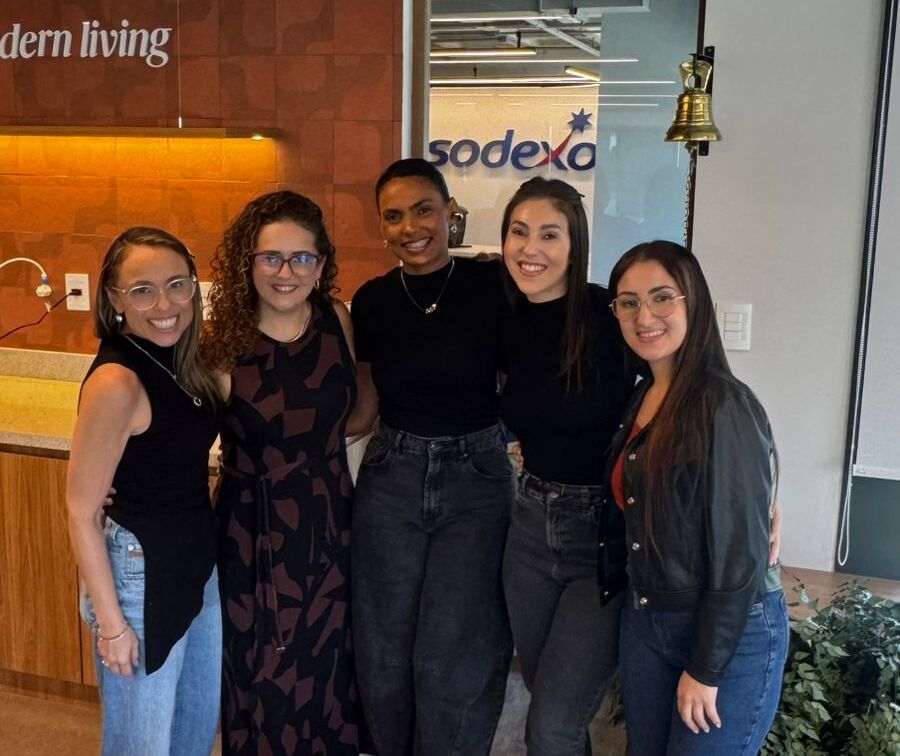 Karina Pavani posa sorrindo ao lado de quatro colegas em um ambiente corporativo da Sodexo. Ao fundo, aparecem a marca da empresa em uma porta de vidro, um sino e elementos de decoração do escritório. 