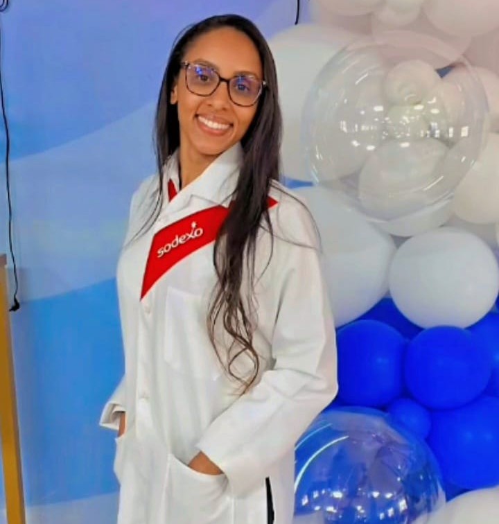 Colaboradora da Sodexo sorrindo, usando jaleco branco com o logo da empresa. Ao fundo, há uma decoração com balões brancos e azuis.