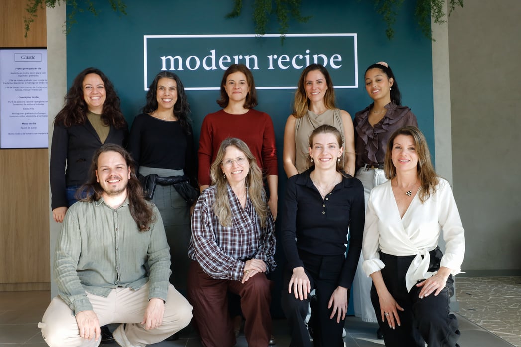  Grupo de nove pessoas posa em frente a uma parede verde com o letreiro “modern recipe”. A imagem mostra um ambiente interno de restaurante, com painel de cardápio ao lado e decoração com plantas suspensas.
