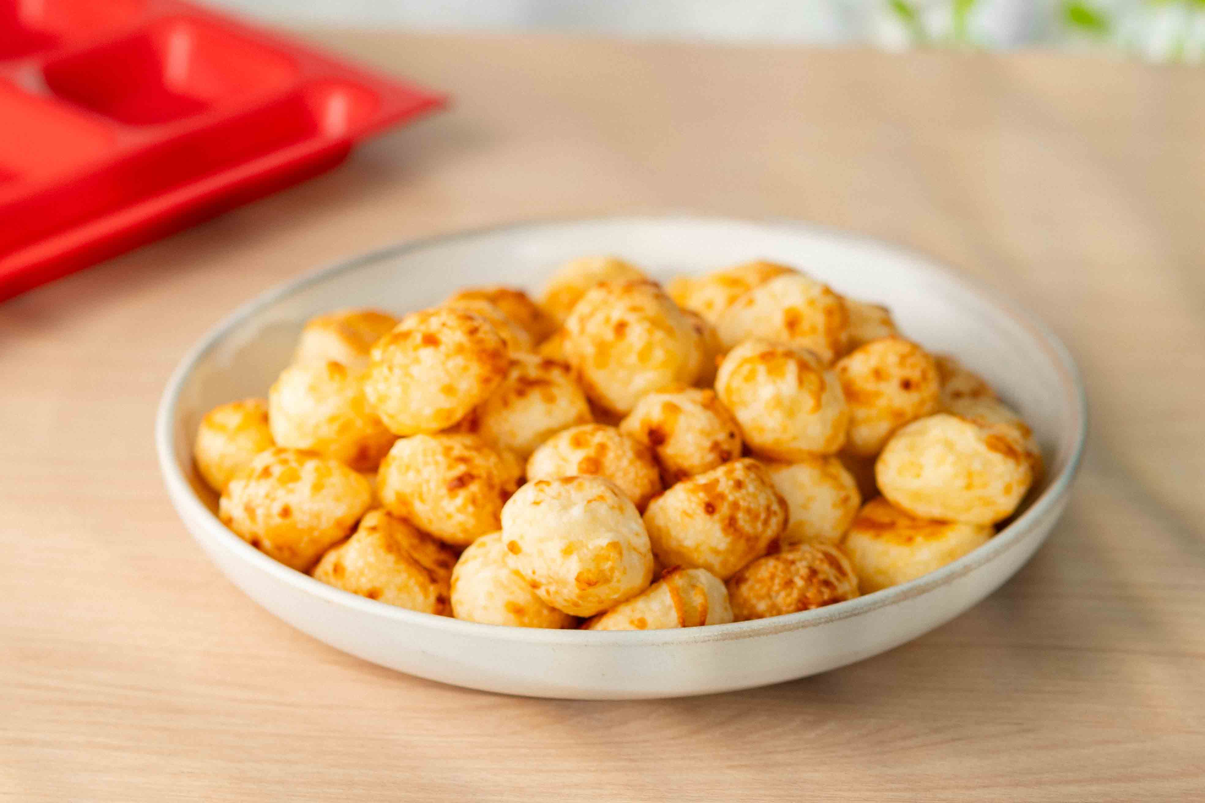 Prato com croquetes empanados dourados dispostos em círculo sobre uma mesa de madeira; ao fundo, tomates-cereja, uma cabeça de alho e alguns dentes de alho desfocados.