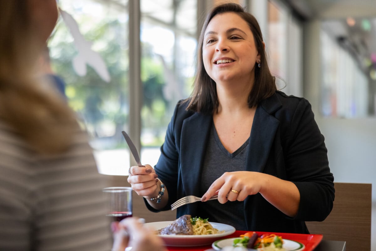 Mulher sorridente, usando blazer escuro, sentada em um restaurante com iluminação natural, segurando garfo e faca enquanto conversa com outra pessoa à mesa. À sua frente há um prato com carne e massa, salada e um copo de suco.