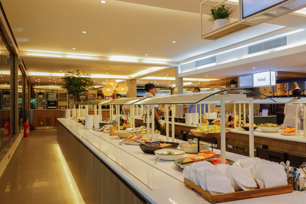 Buffet de restaurante corporativo com saladas expostas, pratos e talheres à disposição, em ambiente iluminado e acolhedor.