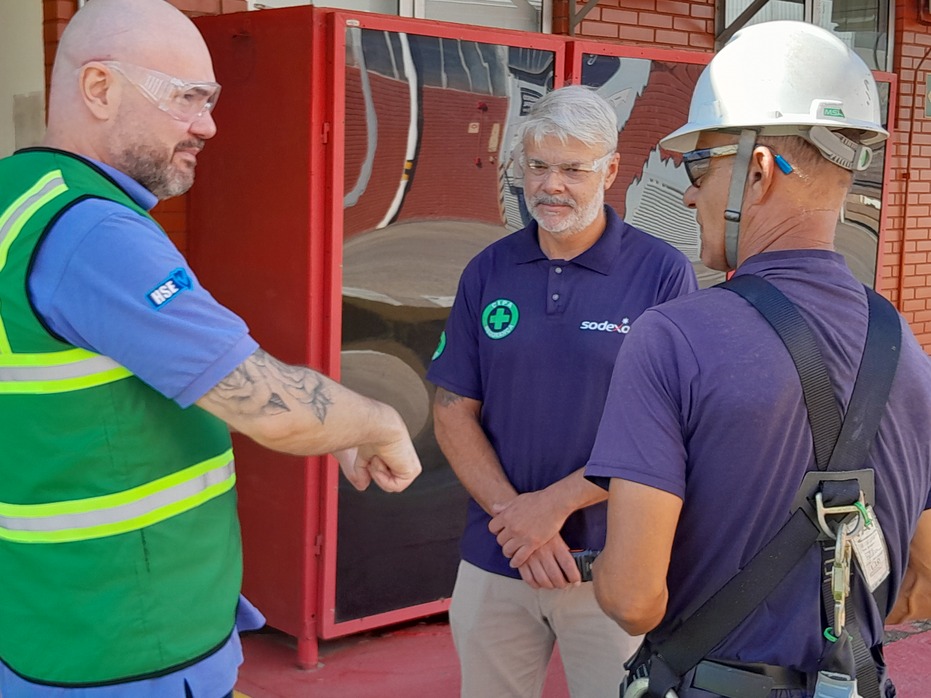  Três profissionais em área industrial realizam um alinhamento de segurança. Um deles, com colete verde de HSE e óculos de proteção, gesticula durante a conversa, enquanto dois colaboradores da Sodexo utilizam capacete e EPIs, atentos às orientações. 
