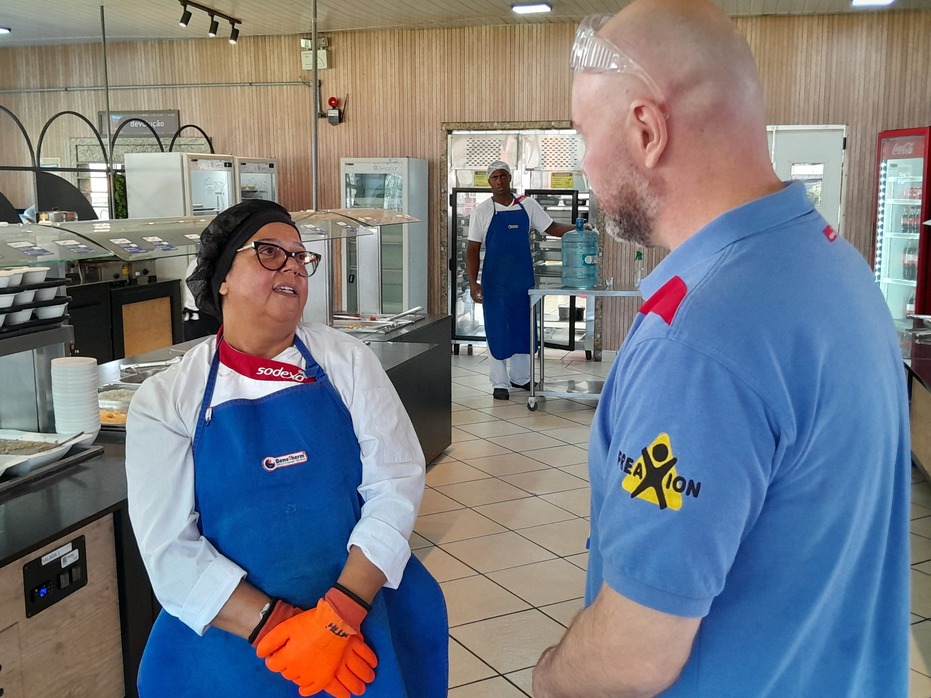 Colaboradora da Sodexo, usando uniforme, touca, luvas e avental, conversa com um profissional em uma área de restaurante industrial. 