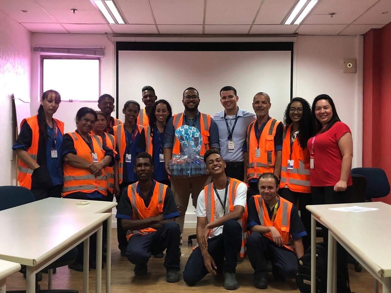 Grupo de pessoas reunidas em uma sala de treinamento, usando coletes de segurança laranja sobre uniformes azuis. Ao fundo, há uma tela de projeção branca e mesas dispostas nas laterais.