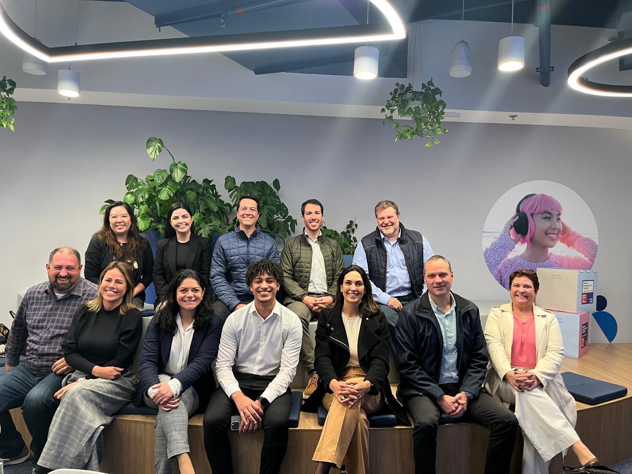 Grupo de pessoas sentado em uma área interna moderna, com plantas decorativas ao fundo. Há uma parede com arte gráfica circular mostrando uma pessoa usando fones. 