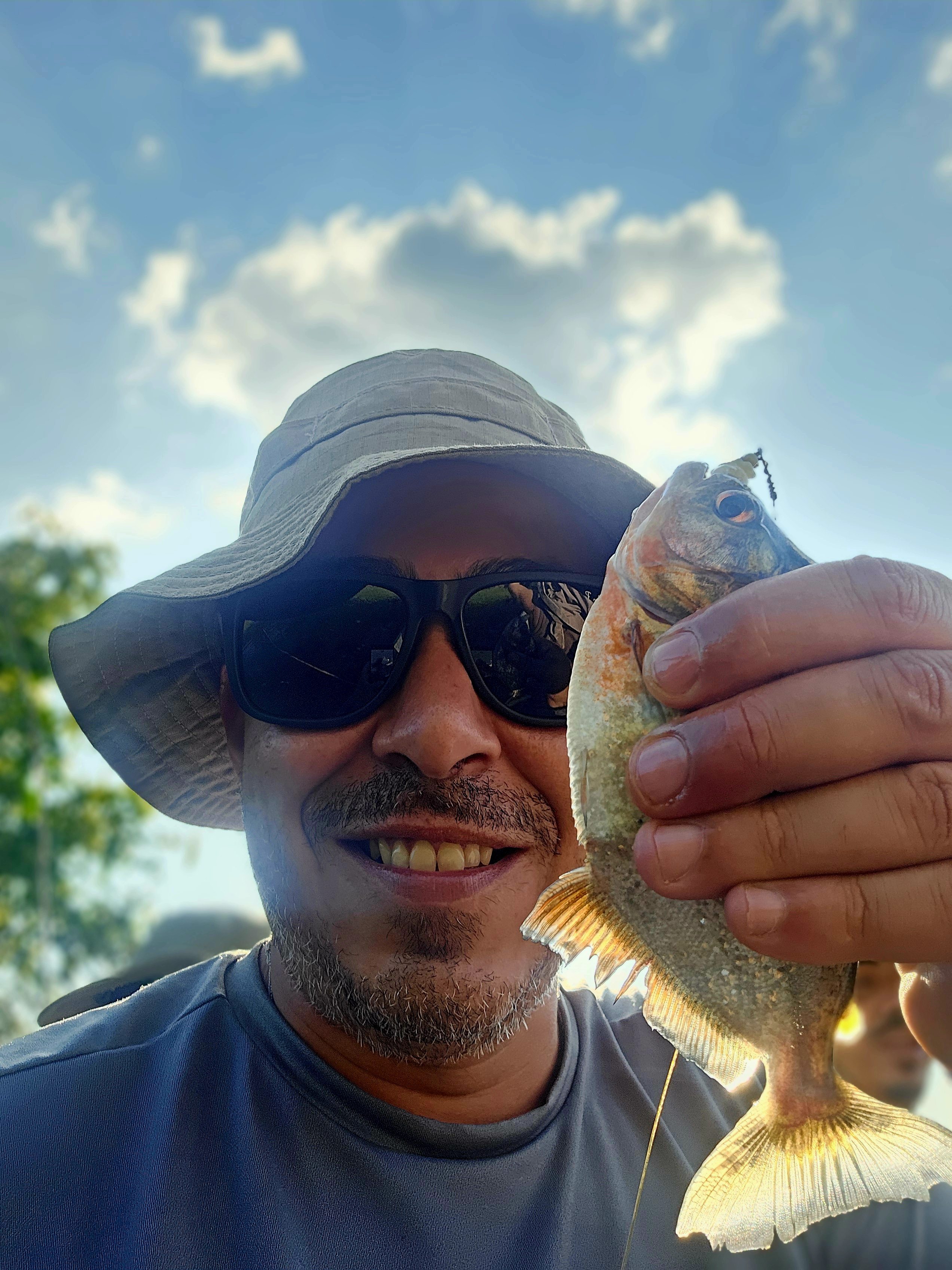 O chef Ricardo Machado sorridente, usa camiseta azul, chapéu e óculos escuros. Ele segura em frente o rosto um peixe que acabou de pescar na Amazônia. No fundo, céu azul e nuvens. 