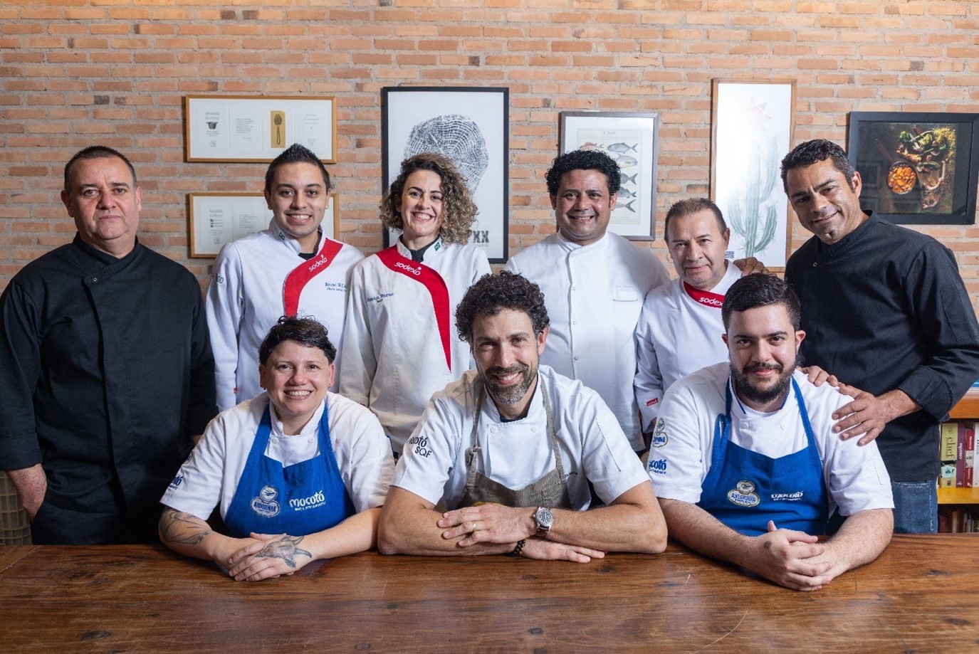 Um grupo de chefs uniformizados posa junto em frente a uma parede de tijolos com fotos emolduradas. Eles estão sorrindo e no centro está o chef Rodrigo Oliveira.