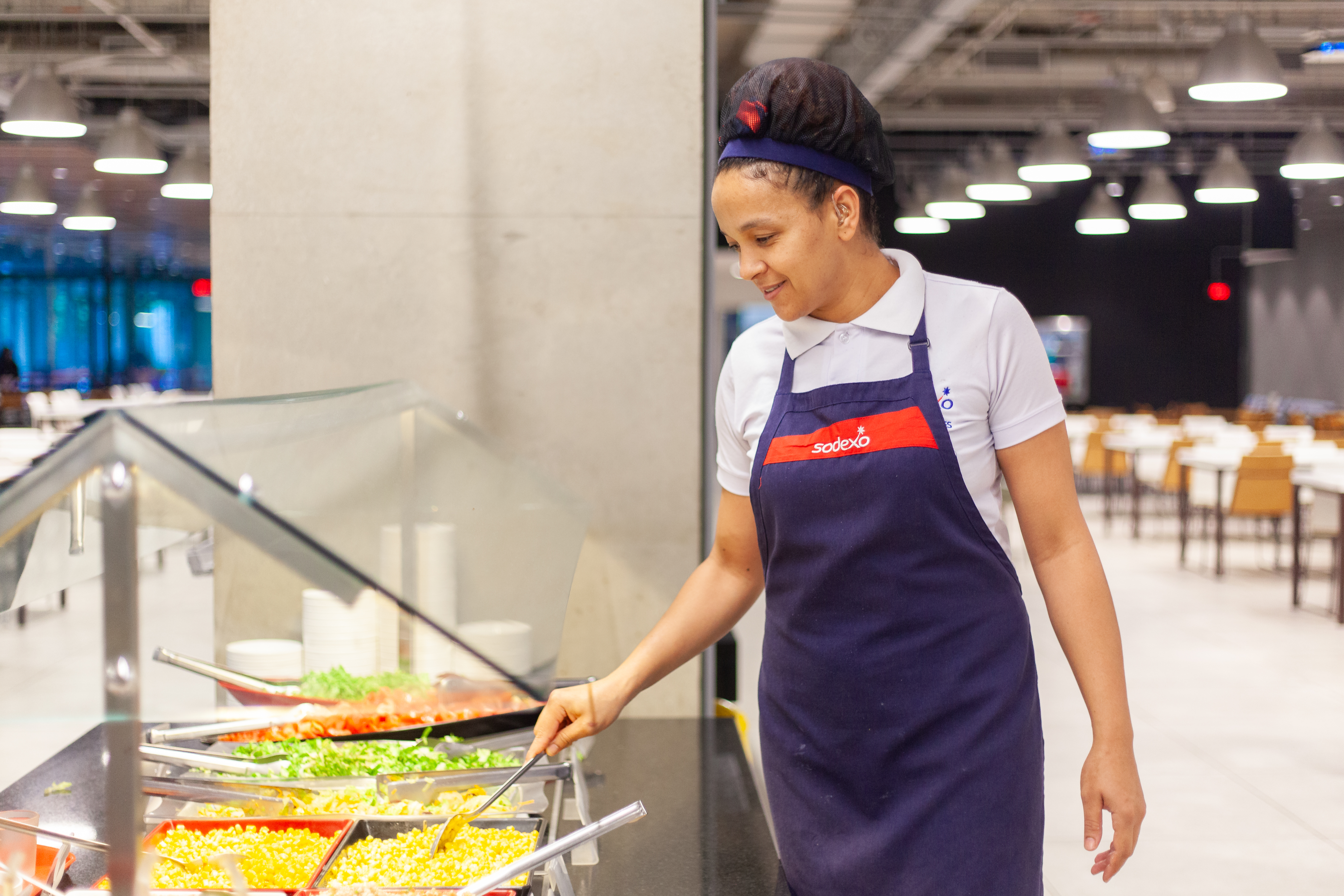 Uma jovem funcionária da Sodexo, com touca e avental azul marinho, sorri enquanto serve comida em um buffet. O buffet possui diversos recipientes com opções de salada, como tomate, milho e outros vegetais coloridos. A cena se passa em um refeitório ou restaurante, com mesas e outros clientes ao fundo, levemente desfocados.