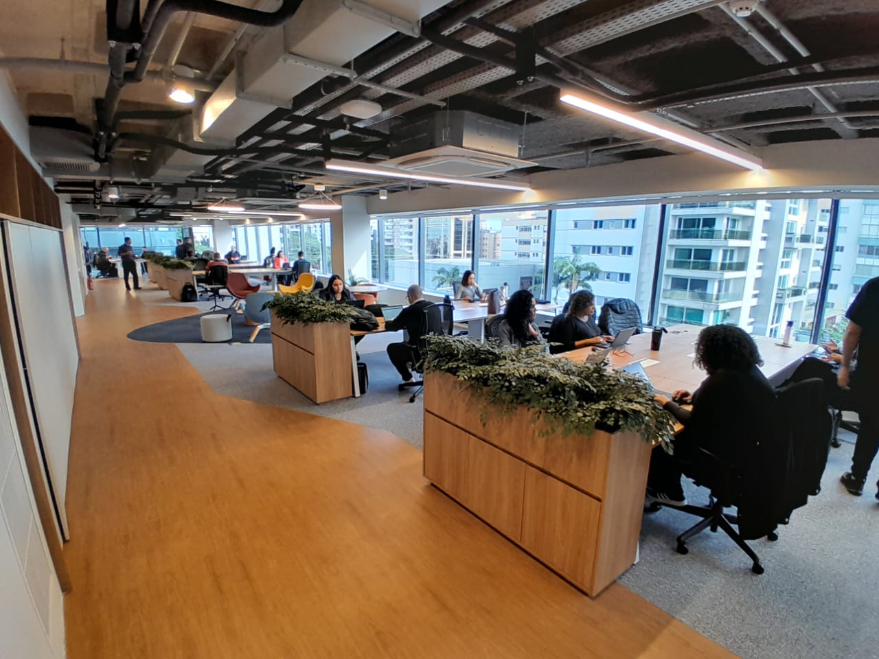 Ambiente de escritório amplo e iluminado, com estações compartilhadas de trabalho, divisórias com plantas e vista para prédios, representando um espaço colaborativo e moderno.