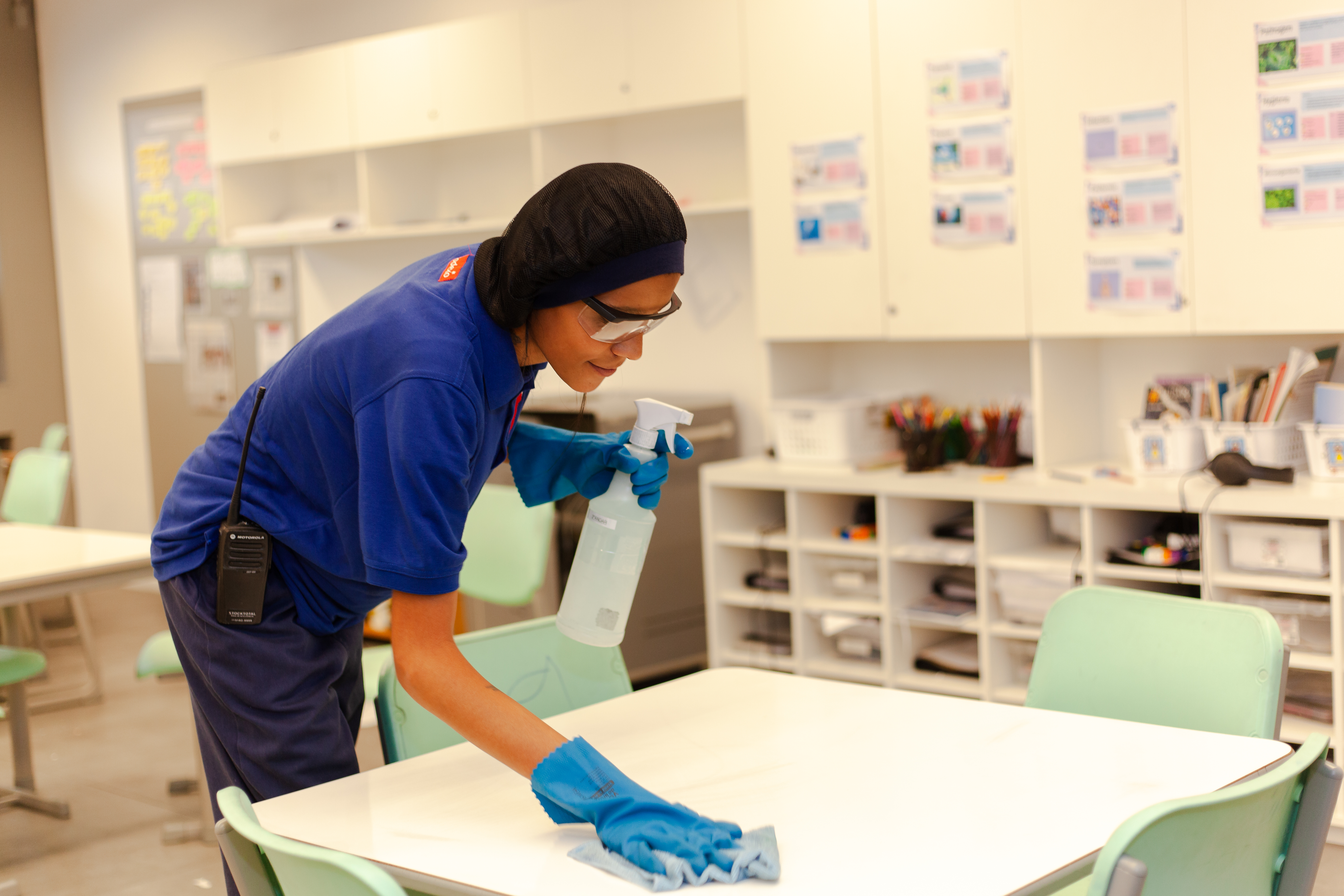 Uma funcionária de limpeza, com uniforme azul, touca, óculos de proteção e luvas azuis, está higienizando uma mesa branca em uma sala de aula. Ela usa um pano azul e um borrifador com produto de limpeza. A sala possui mesas e cadeiras verdes e prateleiras ao fundo. A funcionária usa um rádio comunicador em seu cinto, indicando que ela pode estar em contato com outros membros da equipe de limpeza. O ambiente está limpo e organizado, transmitindo uma sensação de cuidado e higiene.