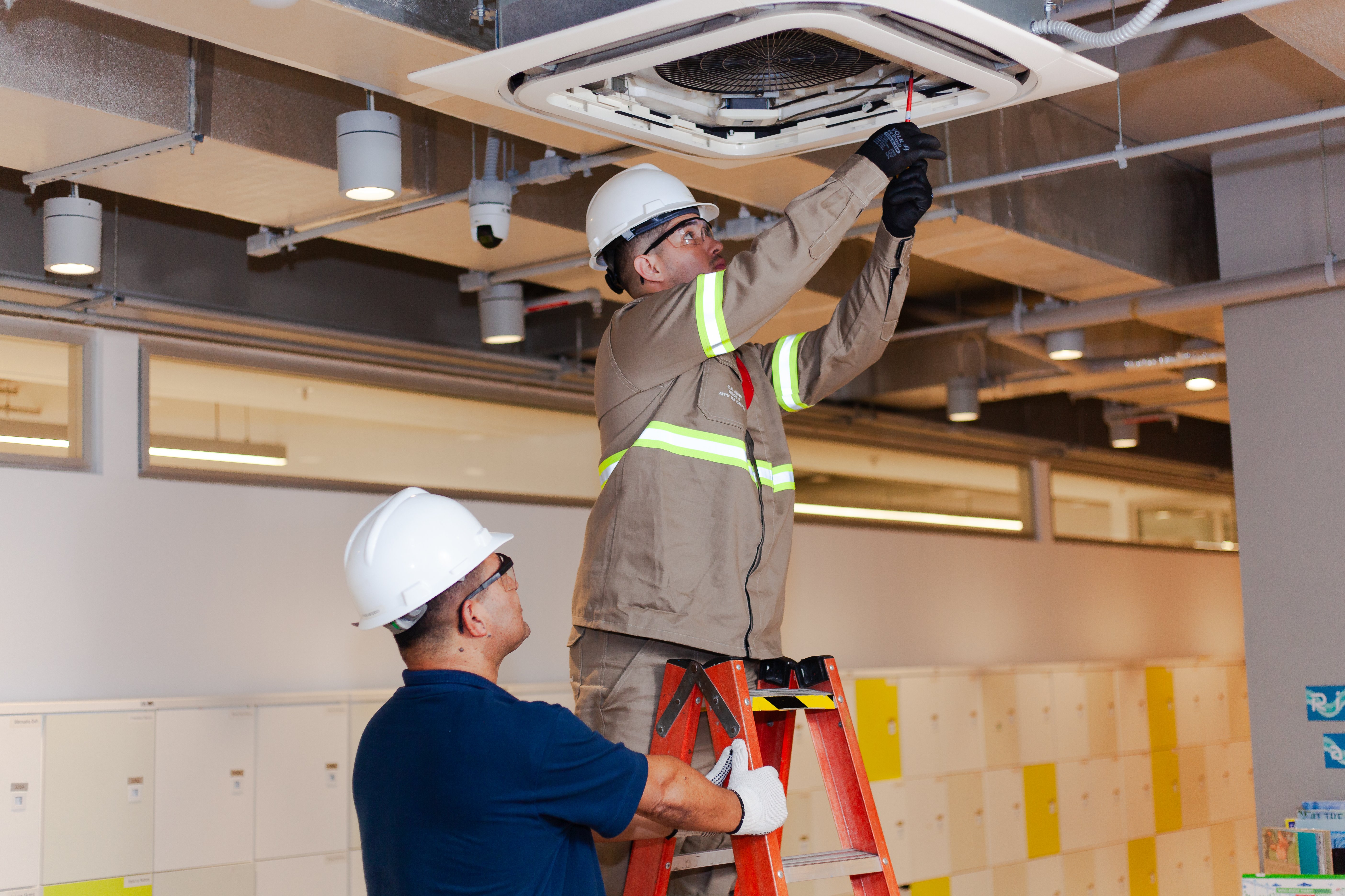 Dois técnicos realizam a manutenção de um sistema de ar-condicionado instalado no teto de uma escola. Um técnico, vestindo uniforme bege com detalhes refletivos e capacete branco, está em pé em uma escada vermelha, utilizando uma ferramenta para realizar ajustes no equipamento. O outro técnico, com camisa azul marinho e capacete branco, está no chão, segurando a escada e observando atentamente o trabalho do colega.