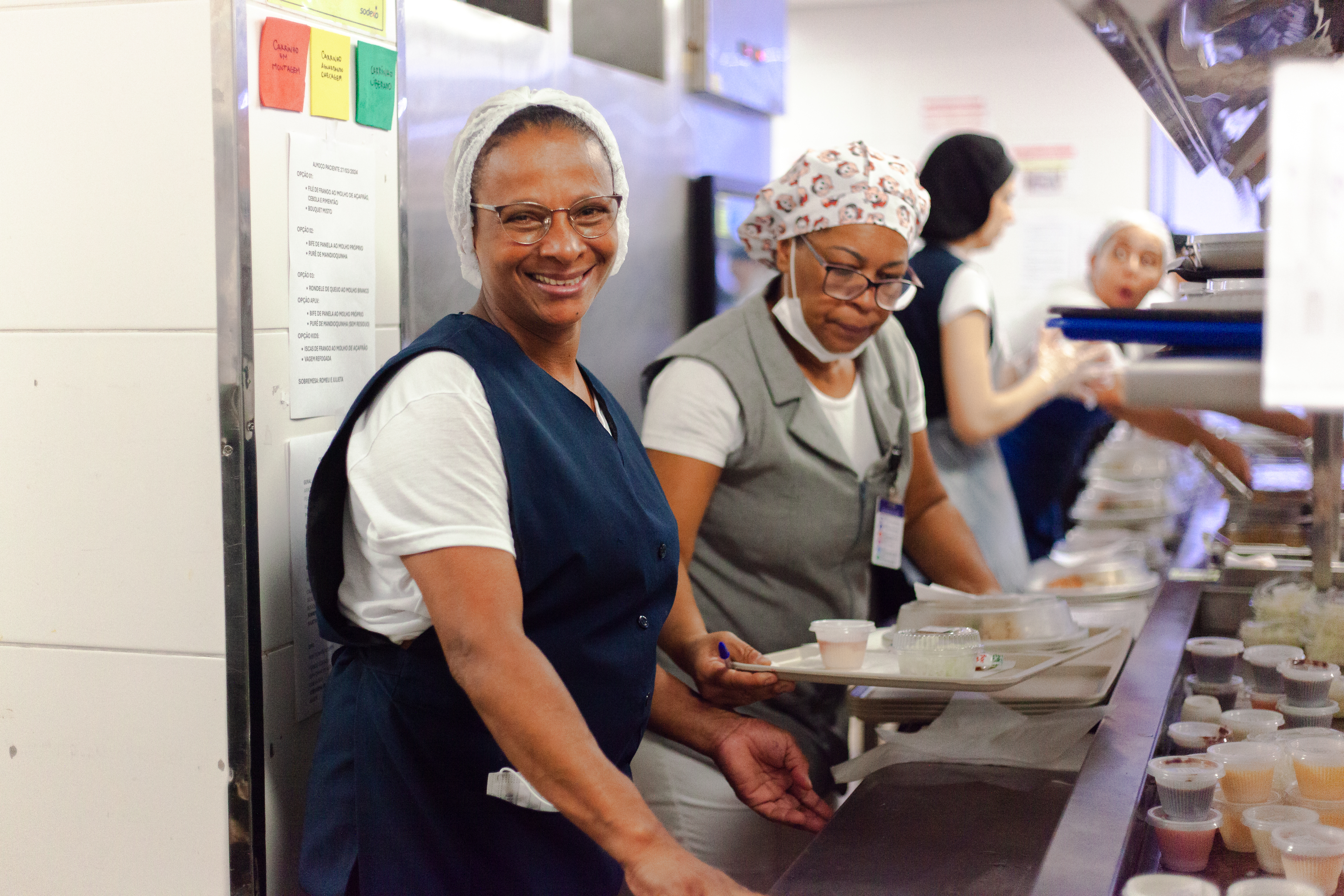 Colaboradoras da Sodexo atuam no preparo e na organização de refeições em uma cozinha de restaurante corporativo.