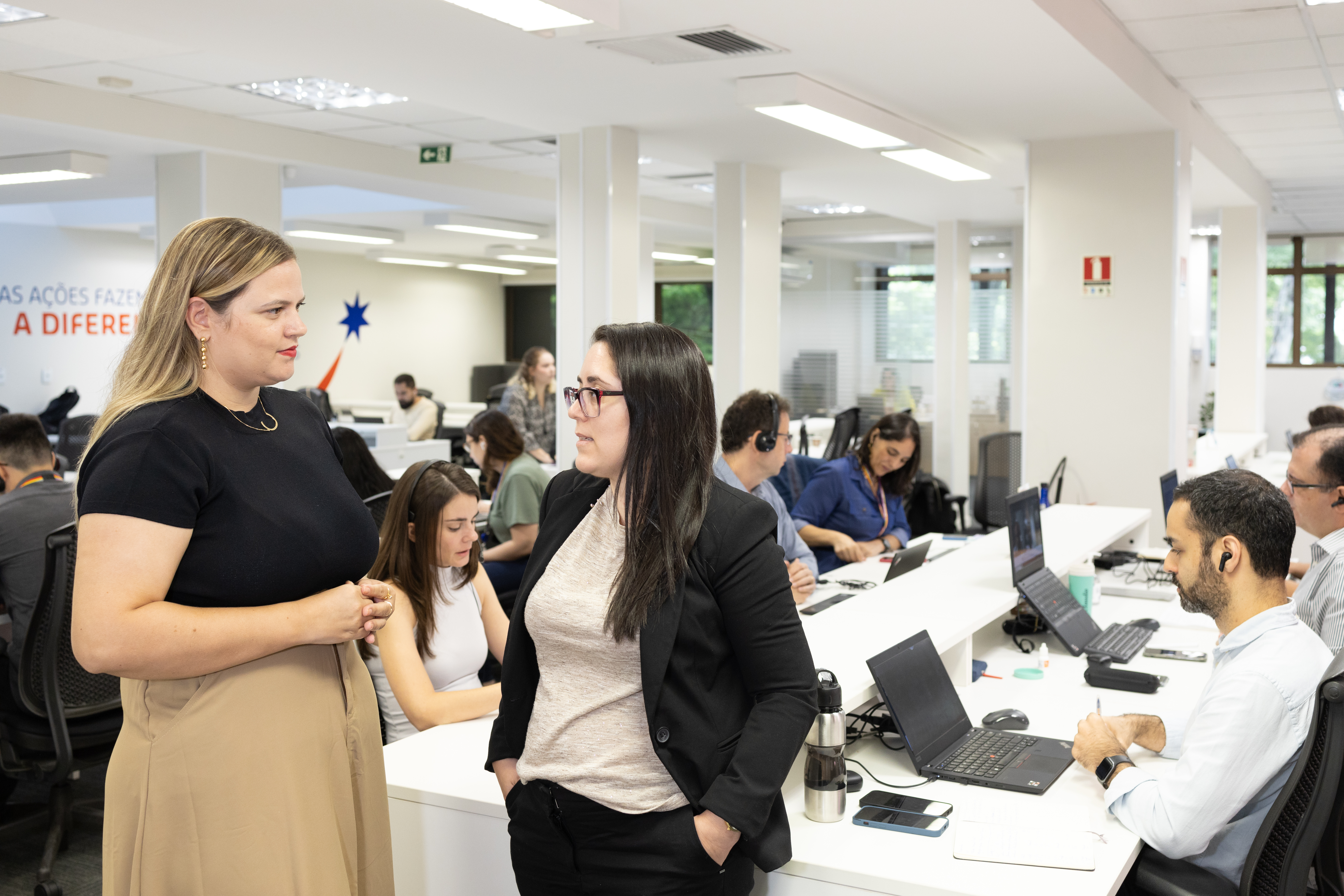 Foto de um escritório amplo e iluminado, com profissionais trabalhando em computadores e duas mulheres em primeiro plano conversando em pé ao lado das estações de trabalho.