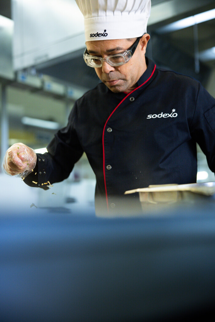 Chef da Sodexo, usando uniforme e touca, prepara prato na cozinha profissional com atenção aos detalhes. 