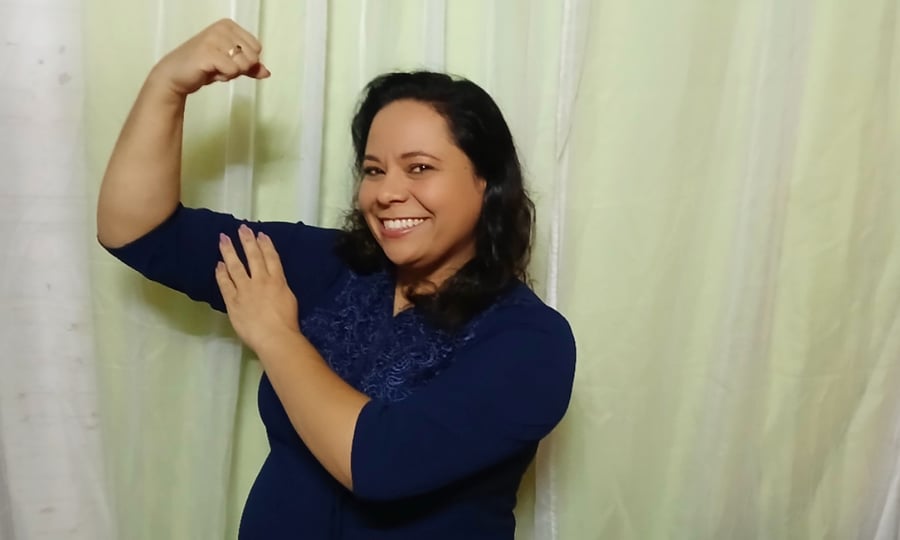 Mulher sorrindo e flexionando um dos braços, vestindo uma blusa azul marinho com detalhes rendados.