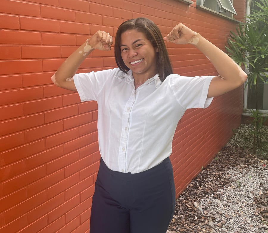 Mulher sorrindo e flexionando o braço em frente a uma parede de tijolos laranjas. Ela veste uma camisa branca com um logo vermelho e calça azul marinho.