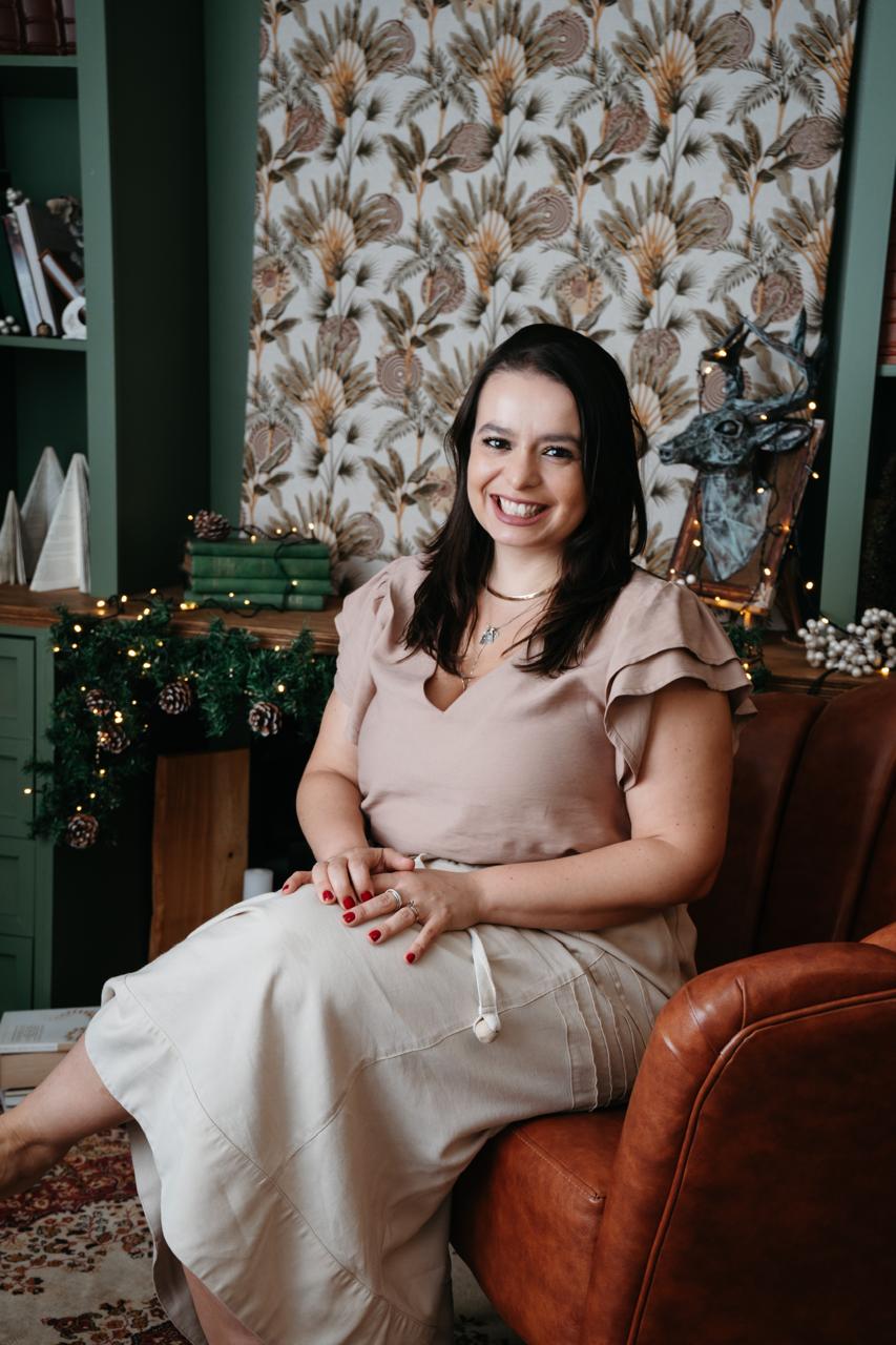Alt: Mulher sentada em uma poltrona marrom, sorrindo para a câmera. Ela veste blusa rosa claro e saia bege, com as mãos apoiadas no colo. Ao fundo, há uma estante, livros, decoração natalina. 