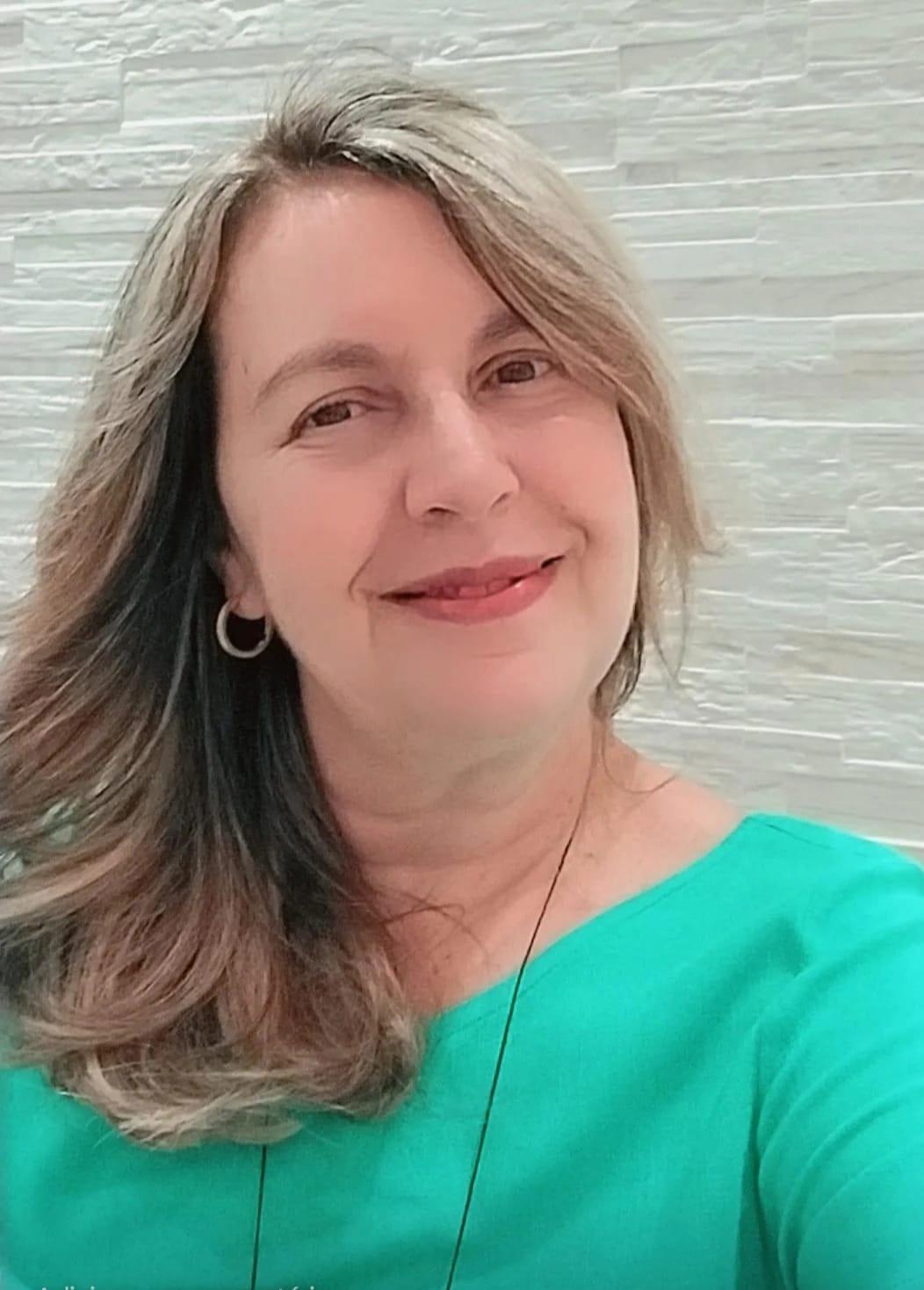 Mulher sorrindo em foto de perfil, com cabelo loiro e liso, usando blusa verde e brinco de argola. Ao fundo, uma parede clara com textura de pedras retangulares. 