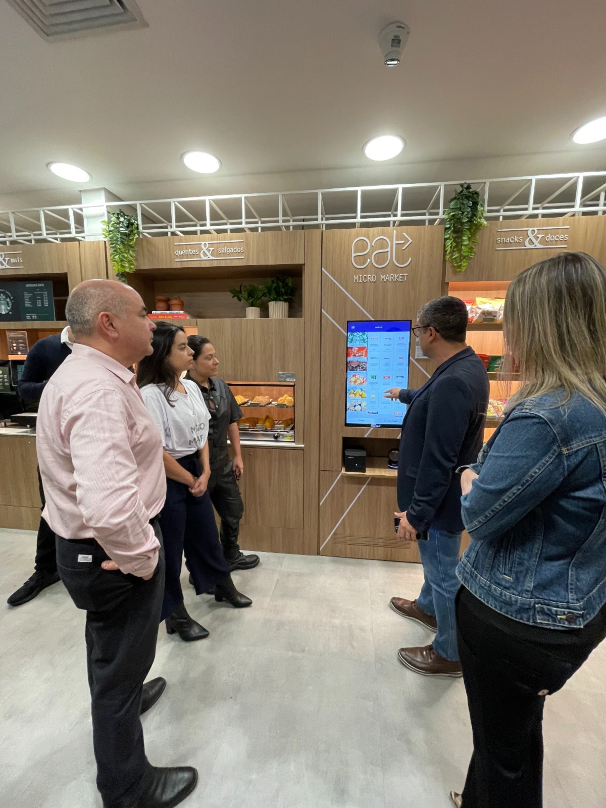 Grupo de pessoas em frente a um micromercado da Sodexo, observando a demonstração de uso de uma tela interativa para seleção e compra de produtos. O ambiente moderno conta com prateleiras organizadas, vegetação decorativa e iluminação aconchegante.