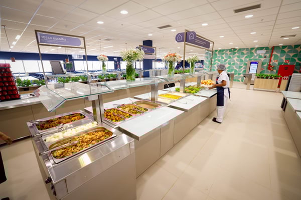 Restaurante moderno da Sodexo com ilhas de buffet organizadas, pratos variados e colaboradora servindo alimentos. O ambiente é decorado com vasos de flores e painéis informativos.