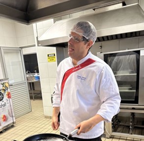 Chef Renan Lemos, da Sodexo, segura uma frigideira enquanto cozinha em uma cozinha industrial. Ele veste uniforme branco com faixa vermelha, touca e óculos de proteção.