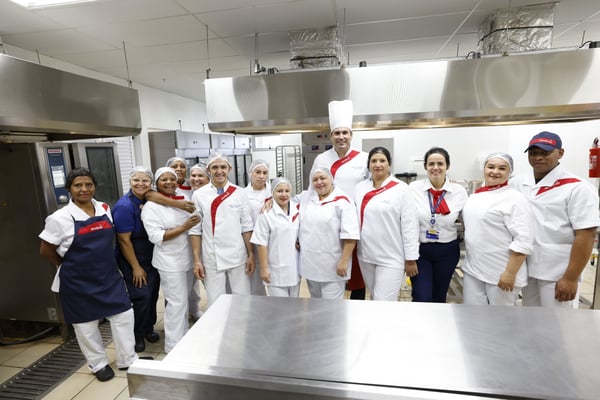 Equipe de colaboradores da Sodexo posa sorrindo em cozinha industrial, vestindo uniformes brancos e toucas de higiene.
