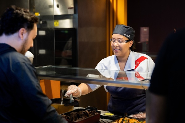 Colaboradora da Sodexo serve uma refeição a um cliente no balcão de um restaurante.
